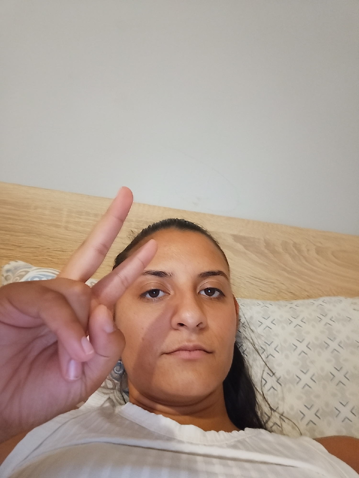 The image is a close-up selfie of a woman lying in bed, holding up two fingers in a peace sign. She has dark hair pulled back and is wearing a white top. Behind her is a wooden headboard and a pillow with a geometric pattern. The background wall is a plain, light color. The lighting suggests an indoor setting, possibly a bedroom. There is no information to determine the location in Yambol, Bulgaria.