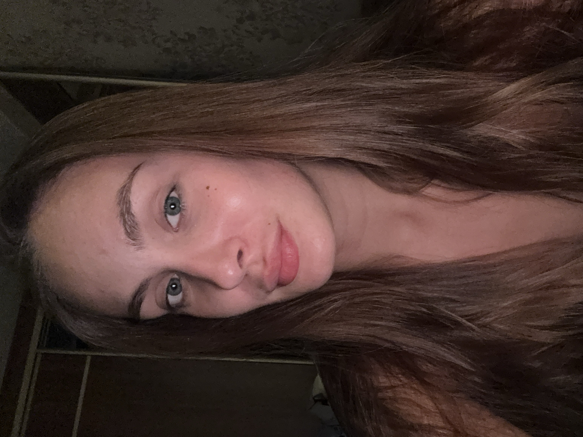 A close-up, frontal selfie of a young woman with long, brown hair. She has light skin with some freckles on her cheeks and nose, and her eyes are a light blue-grey color. Her lips are full and appear to have a subtle pink lipstick. The background is dark and out of focus, with hints of what might be closet doors on the left and a textured wall on the right. The lighting suggests an indoor setting, possibly at night, casting a warm glow on her face. The image does not provide any specific indicators to determine its location within the city of Шолохово, Russia.
