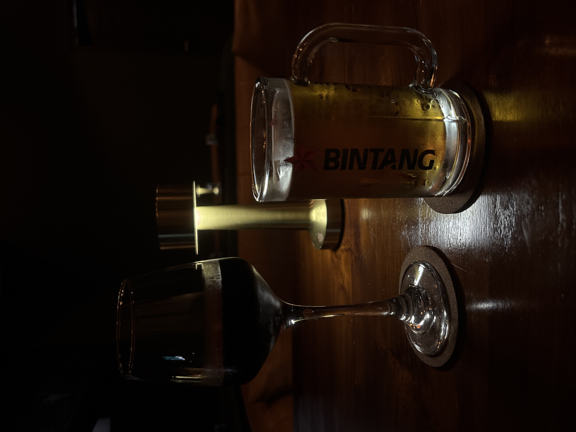 The image depicts a dark, indoor setting, likely a bar or restaurant in Kuta Selatan, Indonesia, based on the presence of a "Bintang" branded beer mug. A beer mug filled with a light-colored beer is resting on a coaster on a dark wooden surface. Next to it is a wine glass, also resting on a coaster, that contains a dark liquid (likely red wine) and a golden doorknob behind both. The scene is dimly lit, with a light source illuminating the objects from above. The focus is primarily on the drinks and their presentation.