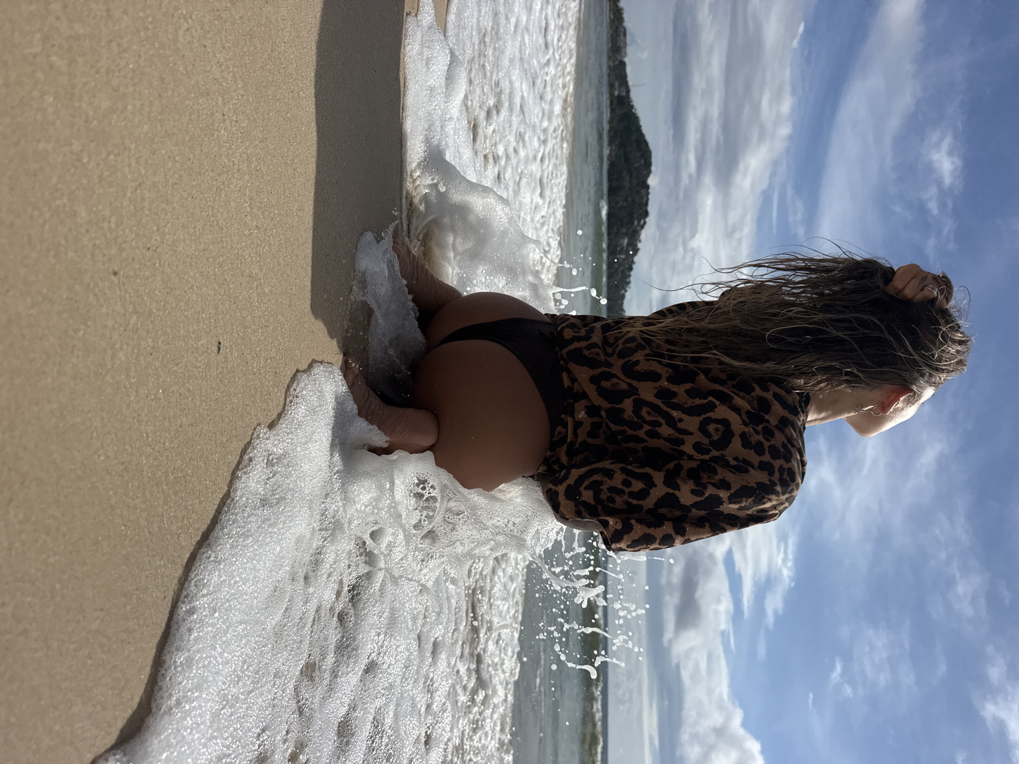 A person is kneeling on a sandy beach in Choeng Thale, Thailand, with waves breaking around them. The person, seen from the back, wears a brown and black leopard-print shirt and dark bikini bottoms. Their long, wet hair is pulled back with one hand, and they are looking up at the sky. The ocean is visible behind them, with a distant island and cloudy sky. The water is creating a splash around the person as the wave recedes.