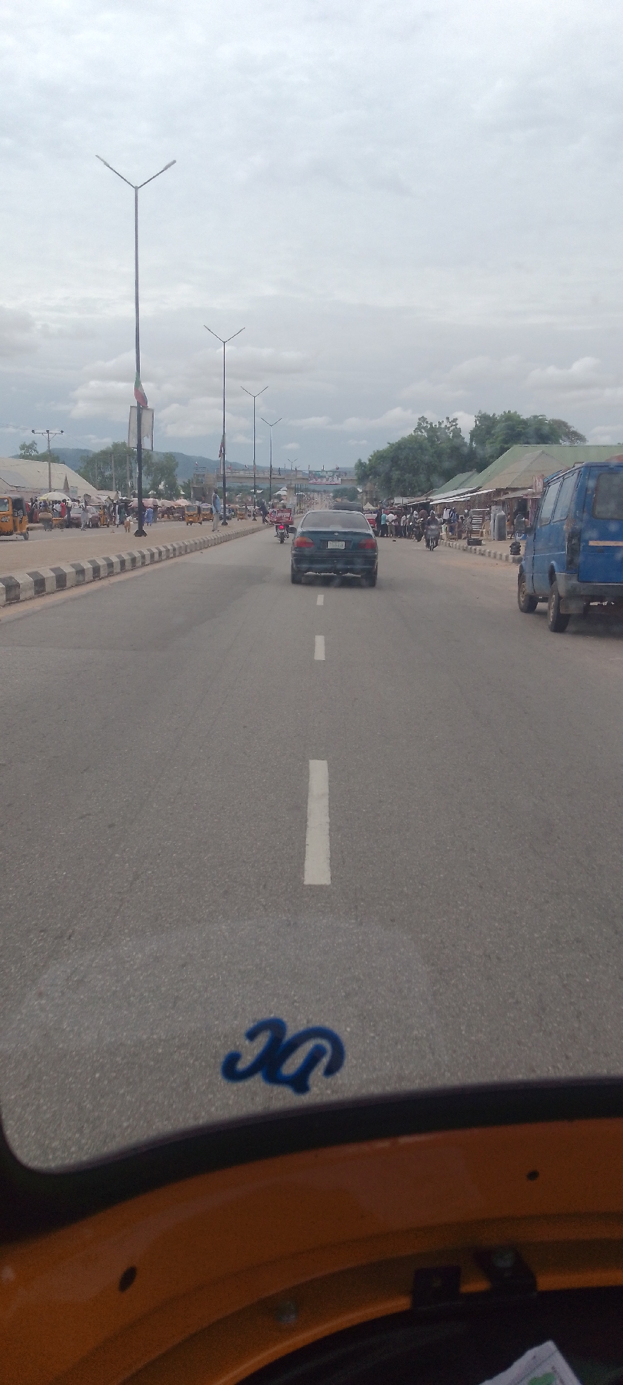 The image shows a street scene in Jalingo, Nigeria, on a cloudy day. The perspective is from a vehicle, possibly a tricycle, looking forward. A black sedan is driving ahead on the asphalt road, which is marked with white lines. On the right side of the road, a blue van is parked, and further down, there are stalls and people lining the roadside. On the left side of the road, there are more stalls, vehicles, and people. Streetlights are visible along the road. In the foreground, a blue emblem is visible on the vehicle's dashboard. The background shows some hills and trees, with buildings and more streetlights suggesting an urban or semi-urban environment.
