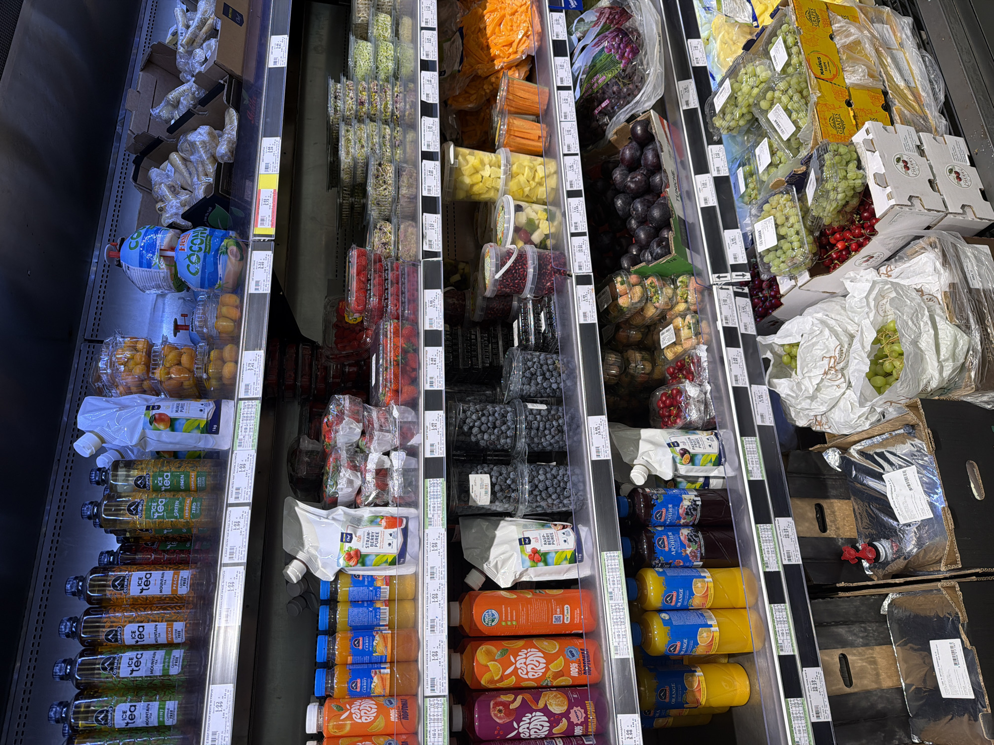 The image captures a refrigerated display case in a supermarket, likely in Burgas, Bulgaria, based on the Cyrillic text on the product labels. The case is stocked with a variety of fresh produce and fruit-based beverages.
On the left side are bottles of "Olympus Ice Tea" in flavors like mint, peach, and green tea. Above them are bottles of coconut water and containers of fresh fruit.
In the middle section are containers filled with cut carrots, pineapple, mixed fruit, pomegranate seeds, and blueberries. There are also pouches of fruit puree, including strawberry and raspberry flavors, as well as bottled carrot and orange juice.
The right section of the display case features various types of grapes (green and red), cherries, and pouches of orange and dark berry juice. A large bag-in-box package of juice is also visible.
The scene is well-lit, typical of supermarket environments. The items are neatly arranged for customer selection.