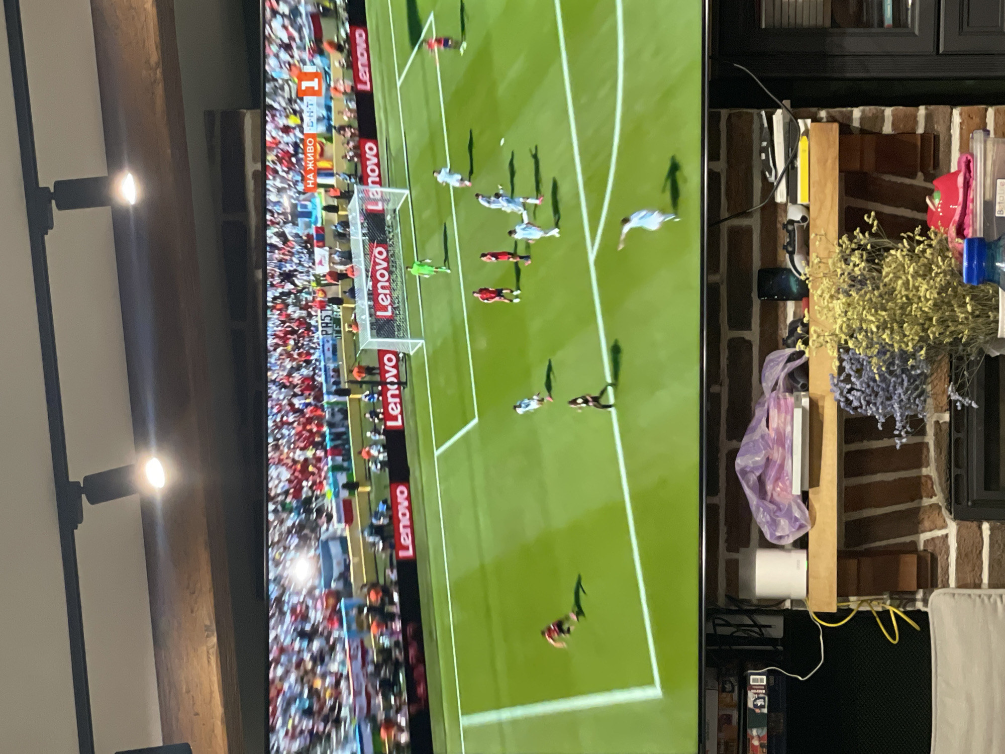 The image shows a television screen displaying a live soccer match. The game appears to be in progress, with players on the field and spectators in the stands. The scoreboard shows "НА ЖИВО" (live) and a score of 1-1. The screen also displays "Lenovo" advertisements. The television is mounted above a brick fireplace, on which several objects are placed, including a white wireless router, a plastic-wrapped package, and dried flowers. A portion of a couch is visible in the lower left corner. The scene is dimly lit by indoor lighting and the bright glow of the television screen. The image was captured in a room in Karlovo, Bulgaria.