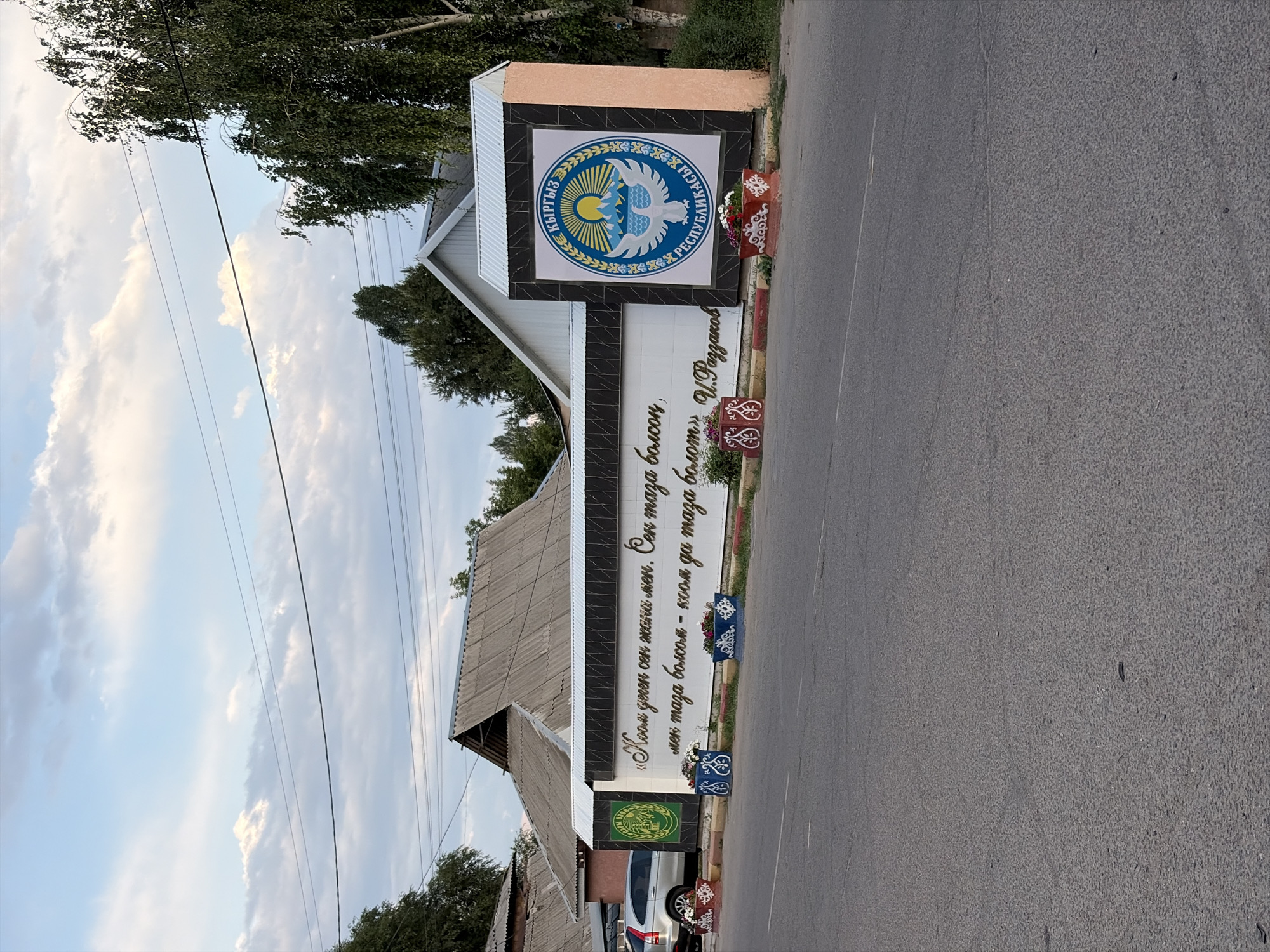 The image captures a street scene in the city of Kant, Kyrgyzstan, featuring a white wall with decorative elements and text. On the left side of the wall, a green emblem with a yellow design is visible. The wall itself displays ornate golden lettering in what appears to be Cyrillic script, along with a larger emblem on the right side. This emblem is circular, predominantly blue and yellow, depicting mountains, a sun, and a bird, with text around its perimeter. Two decorative planters with red and white patterns and filled with flowers are placed in front of the wall, one on the left and one on the right. A silver SUV is parked to the left of the wall, partially visible. The background shows a building with a grey, corrugated roof and trees, set against a sky with scattered clouds. The foreground is dominated by a paved road.