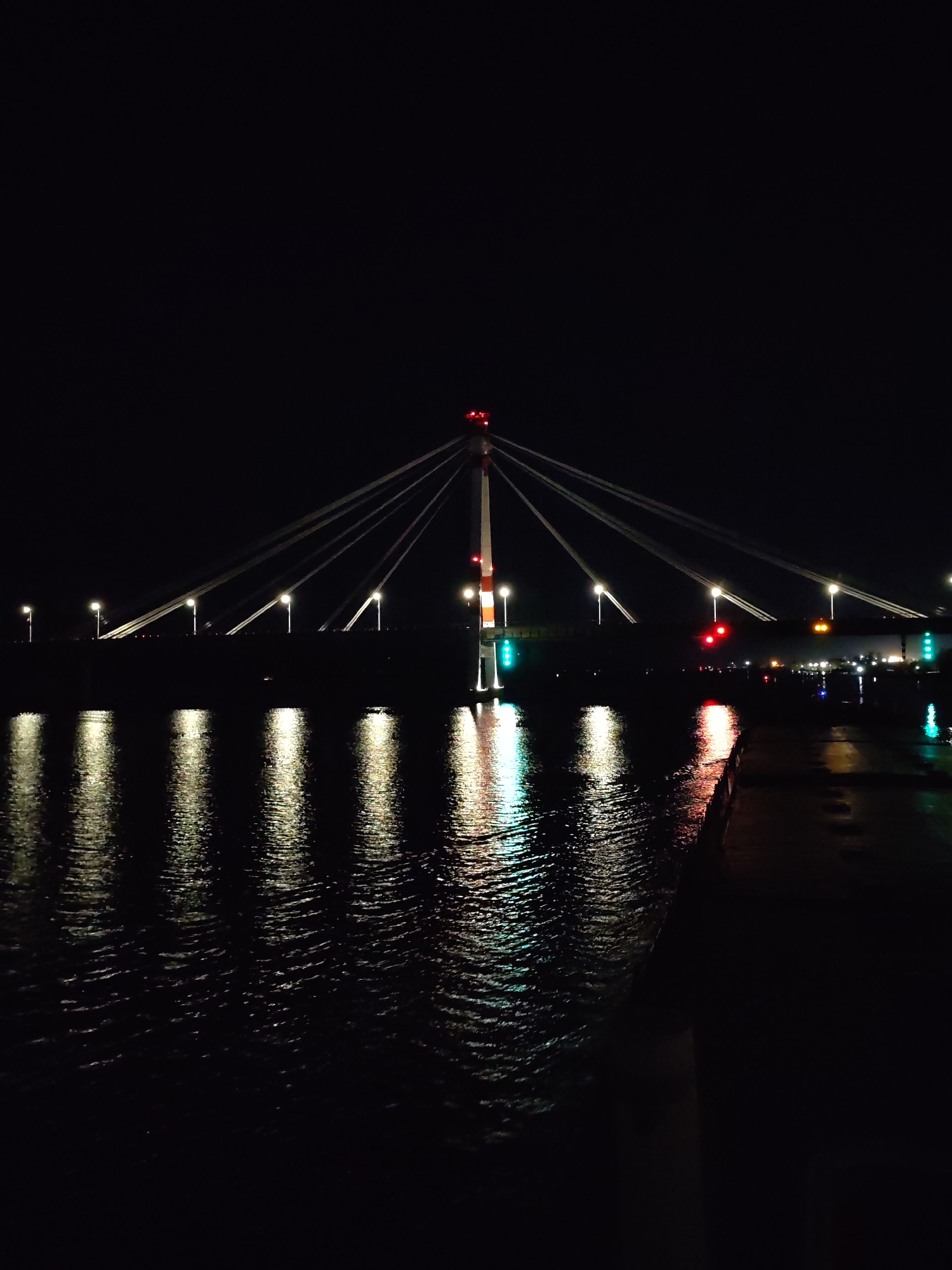 The image depicts a bridge at night in Cherepovets, Russia. The bridge is a cable-stayed structure with a central pylon and multiple cables extending outwards. The water below the bridge reflects the lights, creating vertical streaks of light. There are street lights visible on the bridge deck. The environment is dark, with the focus on the illuminated bridge and its reflections.