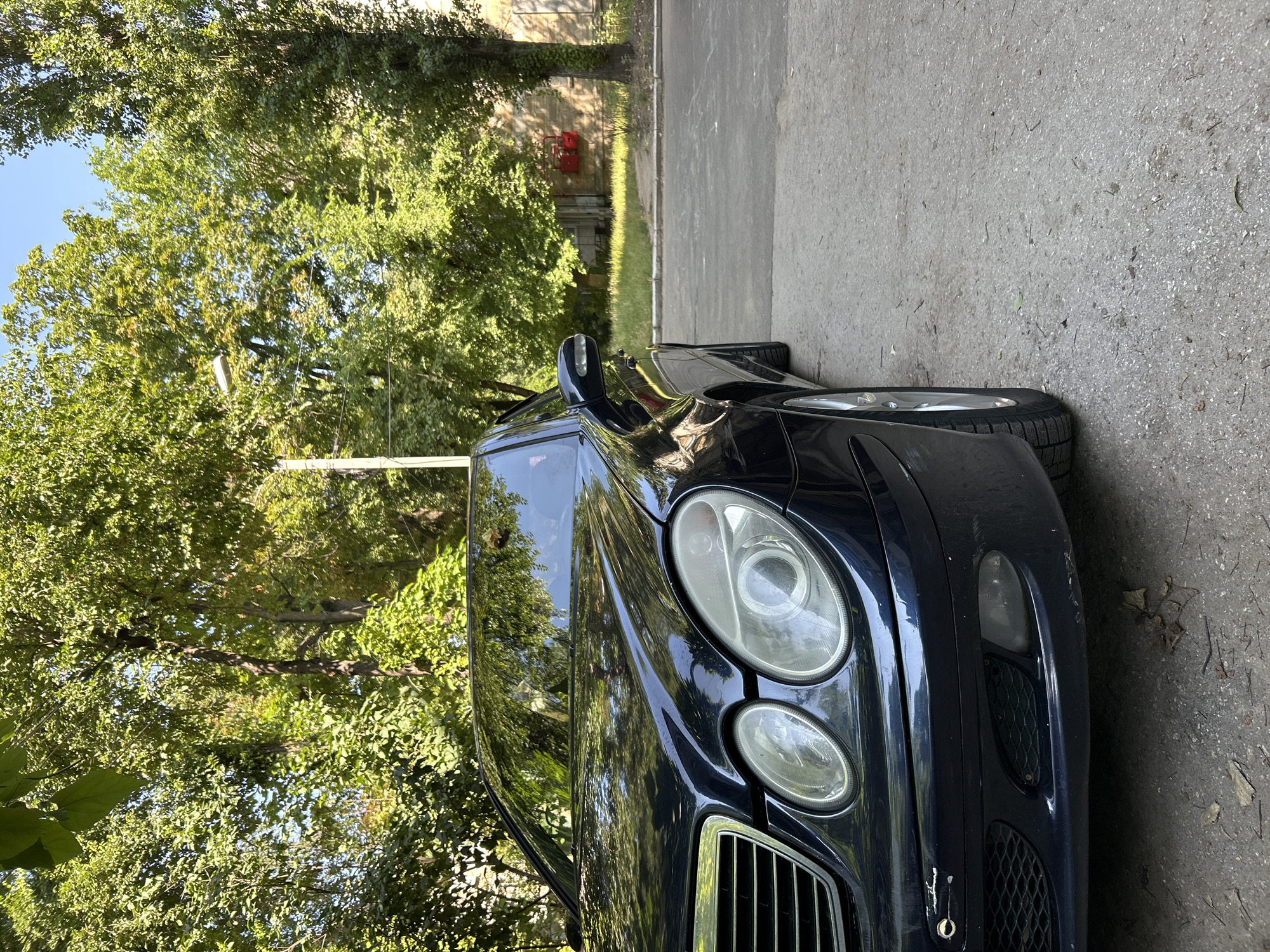 The image shows the front passenger side of a dark blue Mercedes-Benz E-Class sedan parked on an asphalt surface next to a curb. The car's headlights, grille, and part of the hood are visible. Lush green trees with dense foliage form a backdrop, suggesting an outdoor setting, possibly a residential area or park. The sun is shining, casting dappled light and shadows through the leaves. In the background, a building with red fire extinguishers mounted on its exterior wall is partially visible. The scene appears to be a typical urban environment in Chișinău, Moldova, characterized by residential buildings and abundant greenery. There are no people or animals visible in the image. The car exhibits some minor wear and tear, including small scuffs on the front bumper.