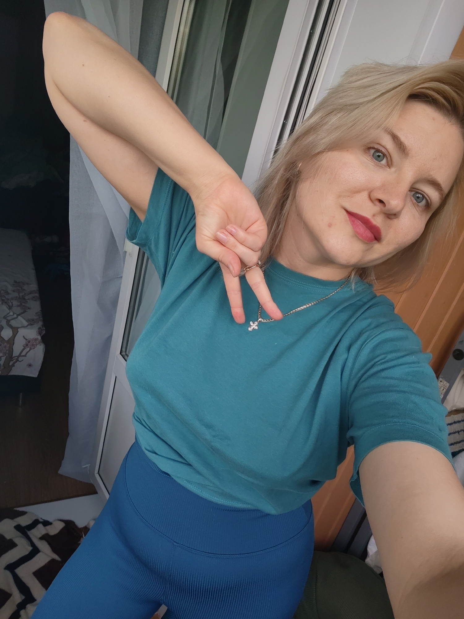 A young woman with blonde hair and blue eyes takes a selfie. She is wearing a teal t-shirt and blue leggings, and has a silver cross necklace on. Her left arm is raised, and she is making a peace sign with her fingers. She has red lipstick on and a subtle smile on her face. The background is blurry, but appears to be an indoor setting with a window and curtains. There is also a bed with patterned bedding visible on the left side of the image. The lighting suggests it is daytime. The woman appears to be in good spirits. There is no visible text in the image.