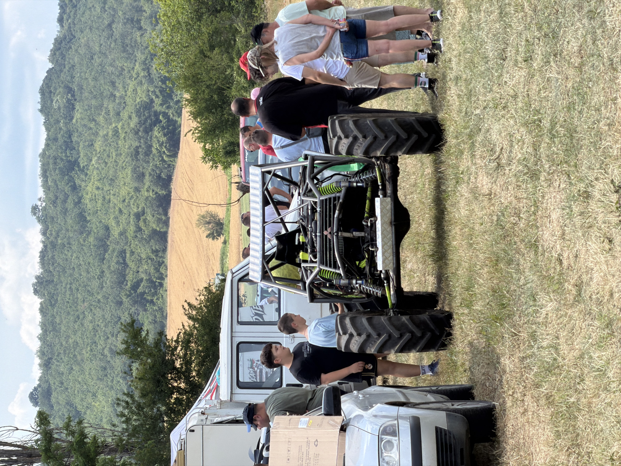 In Bahovitsa, Bulgaria, a group of people stand around a modified vehicle, possibly a monster truck or large off-road vehicle, with prominent knobby tires. The vehicle is brightly colored with a green frame and suspension. Some people are looking at the vehicle, while others are engaged in conversation. The scene appears to be an outdoor event, with a grassy field and a wooded hill in the background. The time of day seems to be daytime, with bright sunlight casting shadows. The weather appears to be fair and warm. The overall atmosphere seems casual and festive, suggesting a community gathering or a car show. There are no visible indications of specific emotions beyond general interest and engagement. The backdrop of rolling hills and greenery suggests a rural or semi-rural location.