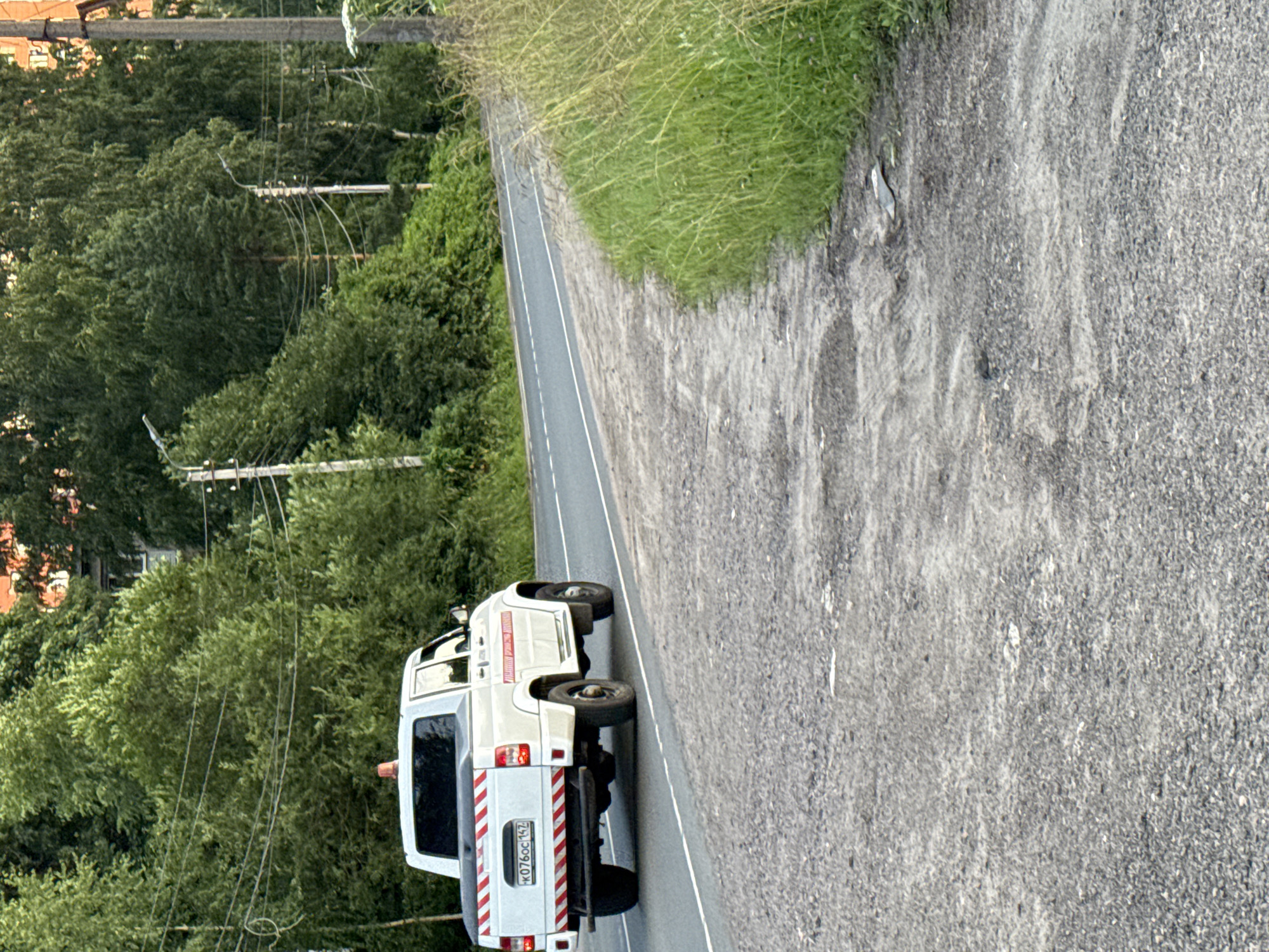 In the city of Pargolovo, Russia, a white UAZ Patriot pickup truck, equipped with an orange flashing light on its roof and red and white reflective stripes on the rear, is parked on the side of a paved road. The truck's license plate reads "K076OC 147". The truck has a red stripe along the side with Cyrillic text which appears to say "дорожная служба" (road service). The road is bordered by a grassy embankment on the right and dense green trees on the left. The environment suggests a rural or suburban setting on a cloudy day, possibly during daylight hours. The road surface is asphalt with visible damage and gravel. There are power lines and utility poles present in the background, amidst the foliage. The truck appears to be stationary, possibly attending to road maintenance or an incident.