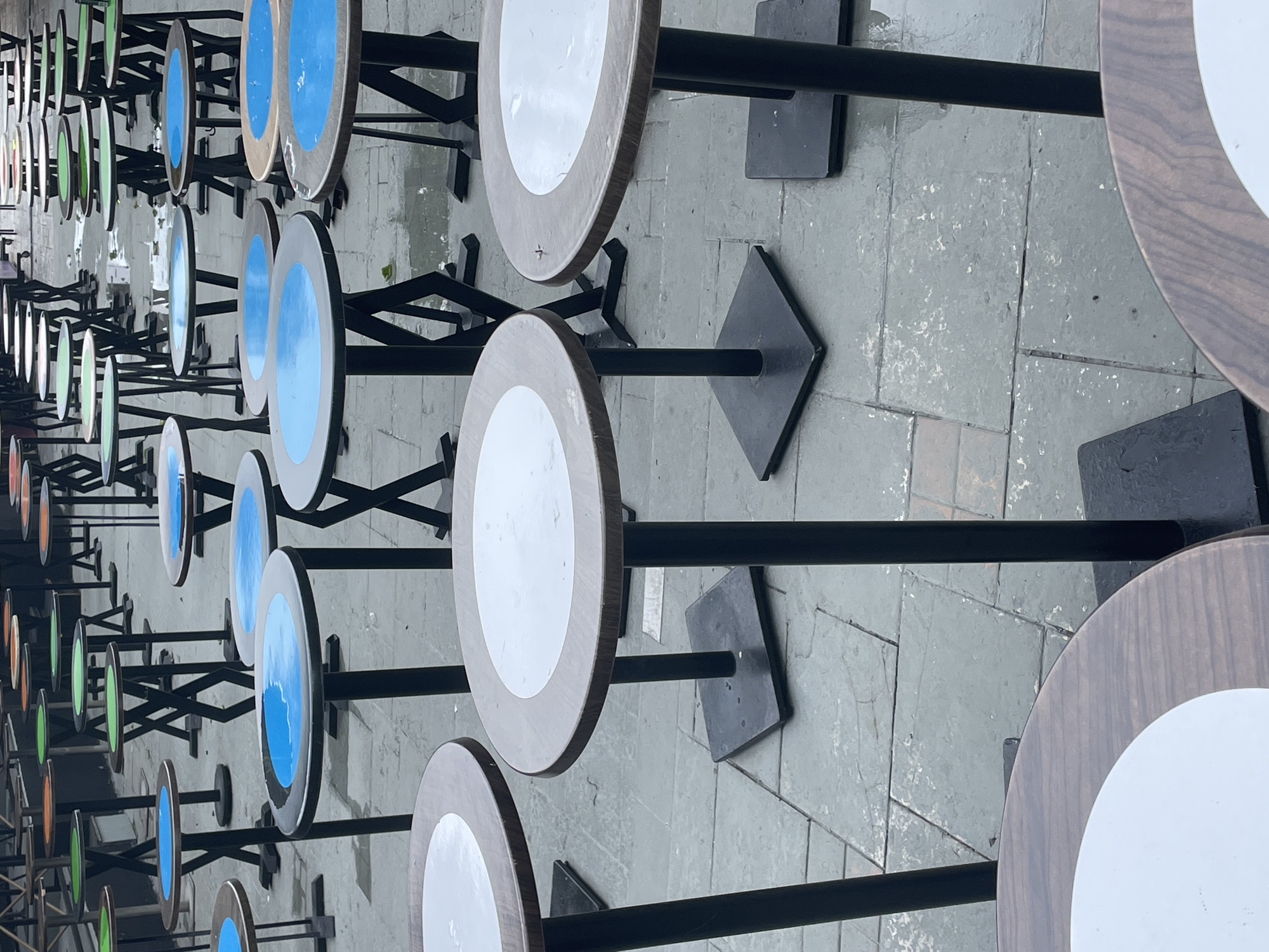 The image displays a series of small, round tables with metal bases arranged outdoors on a paved surface. The tables are lined up in rows receding into the distance. Some tabletops are painted with a blue center, while others are white, and some are painted orange and green. The tables appear to be located outside a restaurant or cafe in Budva, Montenegro. No people are visible in the frame. The focus is on the repeating pattern of tables and their arrangement in an outdoor setting, which suggests they are part of a street cafe or restaurant. The tables feature a brown wood-grain exterior with painted interiors that have contrasting colors. The pavement appears to be stone or concrete tiles. The image has a perspective that makes the tables appear smaller further away.