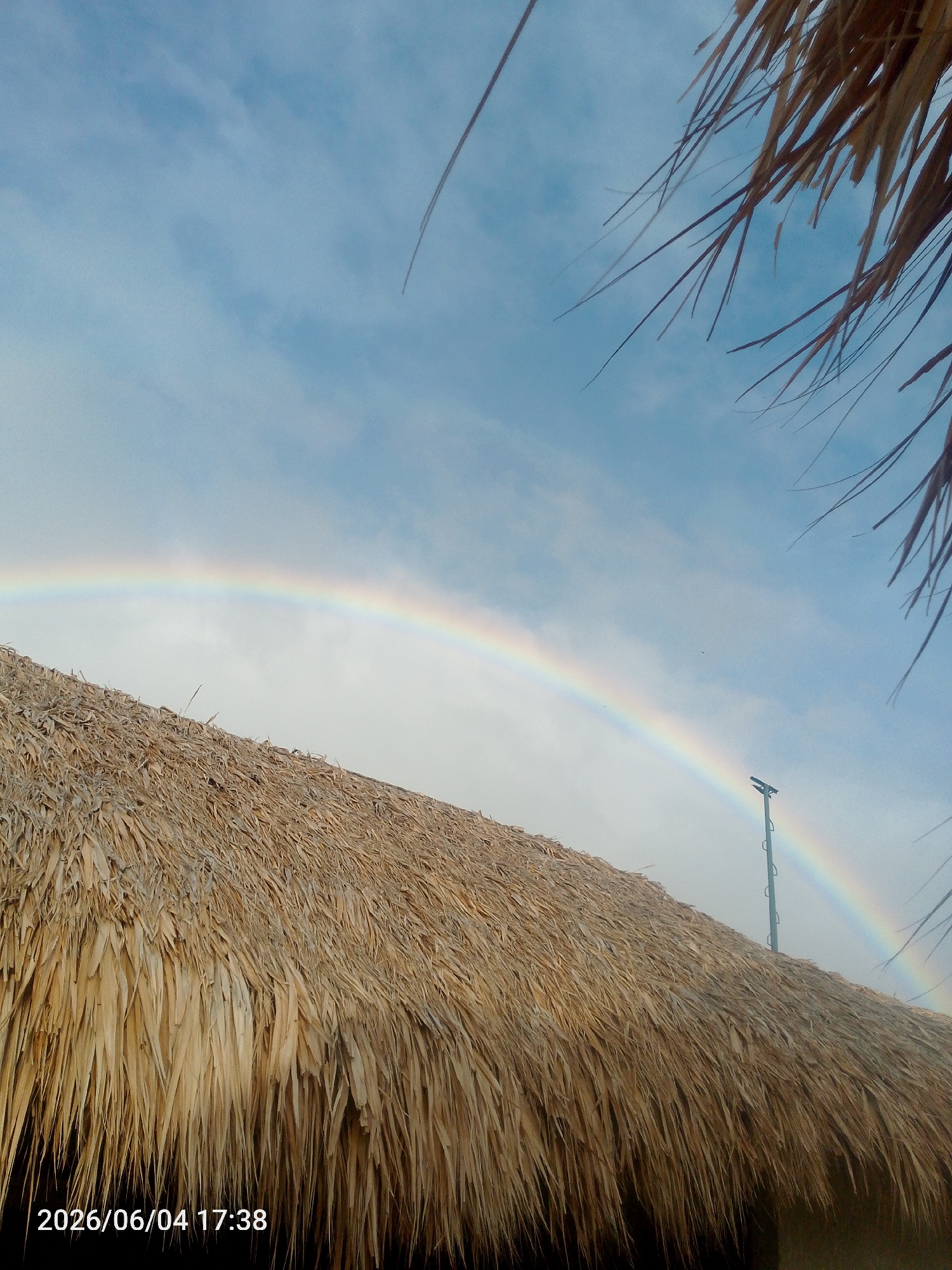 The image displays an outdoor scene featuring a prominent rainbow arching across a partly cloudy, light blue sky. The lower half and left side of the frame are largely occupied by a textured, golden-brown thatch roof made of dried leaves or grass, suggesting a natural or traditional building material. The roof appears to be part of a structure close to the viewer, possibly a hut or a cabana.
In the upper right corner, and subtly in the upper left, dark, thin strands of what appear to be palm fronds or similar dried vegetation are visible, slightly out of focus, implying they are very near the camera. On the right side, behind the rainbow, a tall, slender pole or mast with some attachments stands against the sky. Small, dark specks, possibly birds, are visible against the blue backdrop.
The main event captured is the appearance of a rainbow, indicating recent rainfall or atmospheric conditions conducive to its formation. The sky is largely clear, suggesting the weather is improving. A timestamp in the bottom left corner reads "2026/06/04 17:38," denoting the capture time as late afternoon or early evening. The overall scene, with the thatch roof and potential palm fronds, suggests a tropical or beachside location.