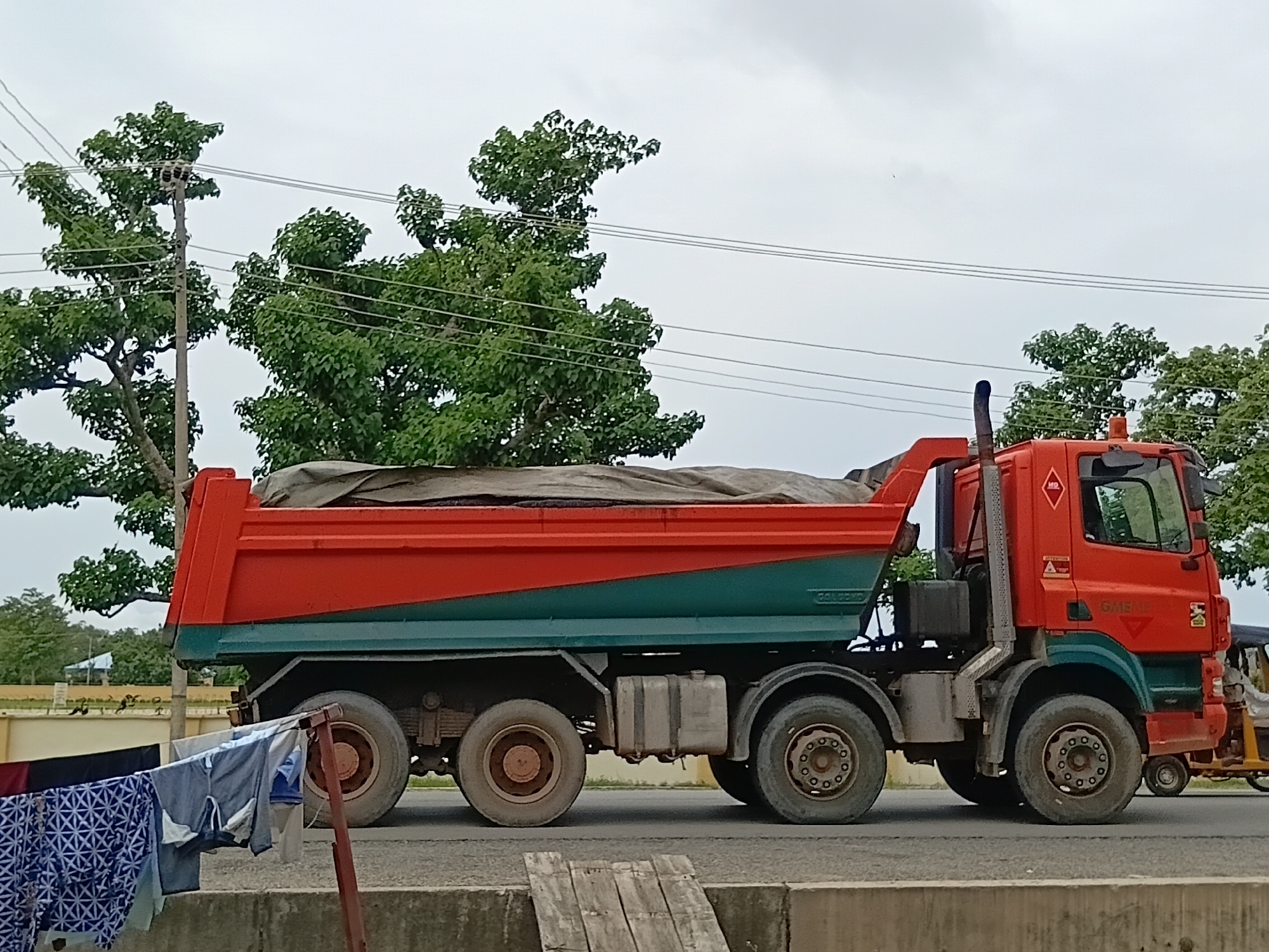 A dump truck, predominantly orange and teal, is parked on a paved road in Jalingo, Nigeria. The truck's bed is covered with a grey tarp, and it is loaded with dark material. Laundry is hanging on a clothesline in the foreground, to the left of the truck. In the background, lush green trees and a cloudy sky are visible. A power pole with wires runs through the trees. The scene appears to be an outdoor setting, likely near a residential area or a roadside where laundry is being dried.