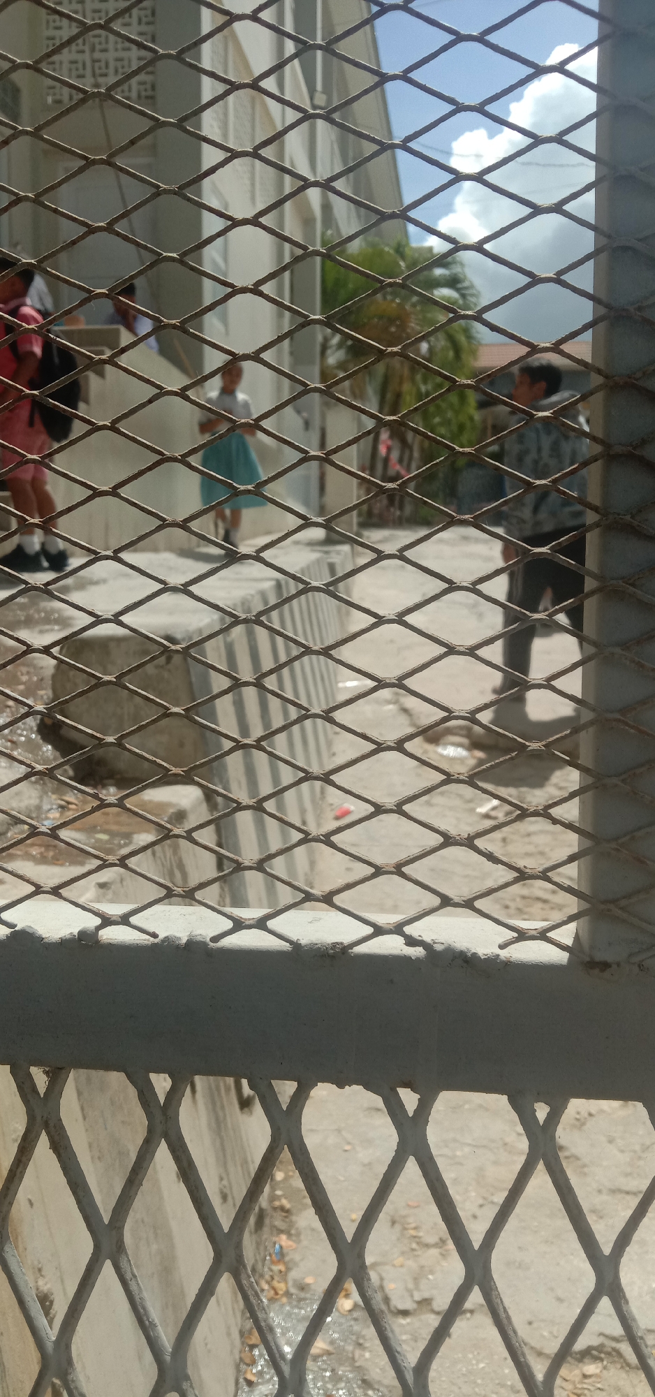 A group of young people are seen standing on a concrete walkway behind a diamond-patterned metal fence. The individuals are blurred, but one appears to be wearing a pink shirt and black shorts, carrying a black backpack. Another person is in a light-colored shirt and a blue skirt. Further back, another individual is visible wearing a patterned jacket and dark pants. The background shows a building with white siding, a palm tree, and a cloudy blue sky. The ground is paved with concrete and there is some debris scattered around. The scene appears to be outdoors in an urban environment, possibly a school or public area. There is no specific indication of the city of Kendari, Indonesia.