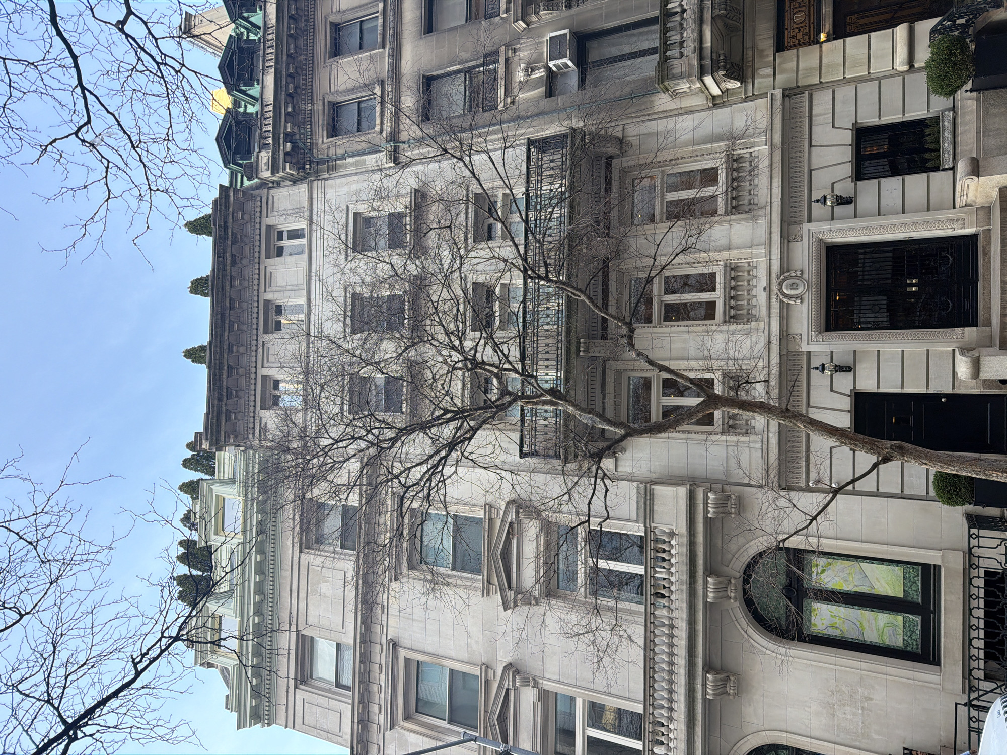 The image displays an upward-angled exterior view of a multi-story townhouse at 13 East 67th Street in Manhattan, New York City, United States of America.
The central building, 13 East 67th Street, is constructed of light-colored stone, exhibiting Beaux-Arts architectural elements. It features a prominent ground-floor entrance with black double doors, an ornate wrought-iron grille, and a carved "13" positioned directly above. Two large, black decorative lanterns flank the main entrance. Multiple rectangular windows with decorative surrounds are arranged symmetrically across its five visible stories, including a prominent balustraded balcony spanning the third floor. The top of the building culminates in a mansard roof with several dormer windows and a row of conical evergreen plants.
To the immediate right, a portion of an adjacent townhouse, constructed of darker grey stone, is visible. It features an exposed window-mounted air conditioning unit on its lower facade. To the immediate left, another light-colored townhouse is partially shown, featuring a large arched ground-floor window containing a multi-panel artwork characterized by green and yellow tones.
A large, leafless deciduous tree dominates the foreground, its bare branches extending across the central building's facade and partially framing the clear, light blue sky. Small green shrubs are visible in planters at ground level. The scene depicts a clear, daylight sky. No individuals, vehicles, or overt activities are present.