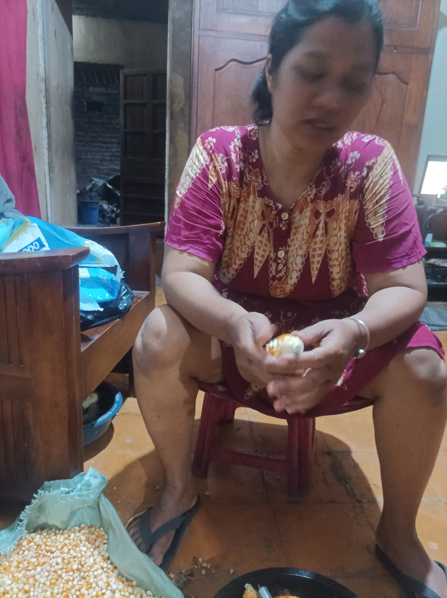 An Indonesian woman, approximately 30-40 years old, is seated on a red stool in a dimly lit room. She is wearing a magenta and tan patterned dress. In her hands, she holds a small, round object, possibly food, which she appears to be peeling or preparing. To her left, a large bag of corn kernels is visible on the floor, and behind her, wooden furniture and a doorway leading to another room are present. The floor is tiled with a warm, earthy tone. The environment suggests a home or a rural setting in Canga'an, Indonesia.