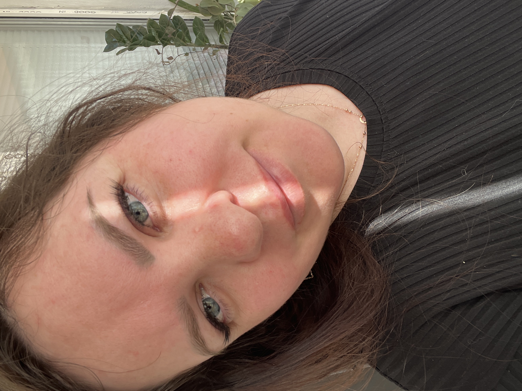 A close-up, rotated portrait shot of a woman with fair skin, light brown hair, and blue-green eyes. She is wearing a black, ribbed top. A delicate gold necklace is visible around her neck. The background shows a window with foliage partially obscuring the view. Location is unknown.