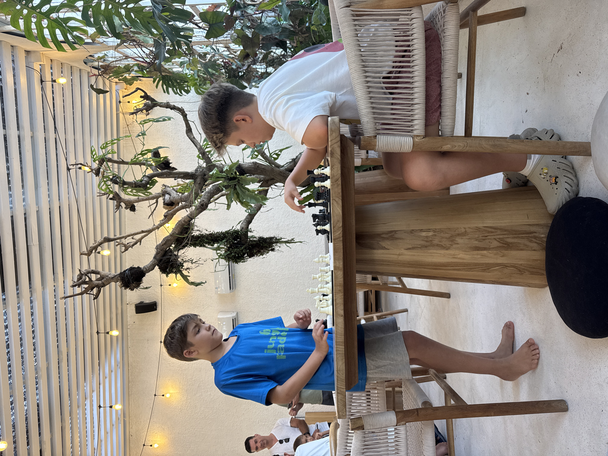 Two boys are playing chess outdoors in what appears to be a restaurant setting. One boy wears a white T-shirt and sits on a woven chair, while the other wears a blue T-shirt and stands barefoot on the ground. The chessboard is set up on a wooden table between them. In the background are plants, string lights, and parts of other tables and people. The location is in North Kuta, Indonesia.