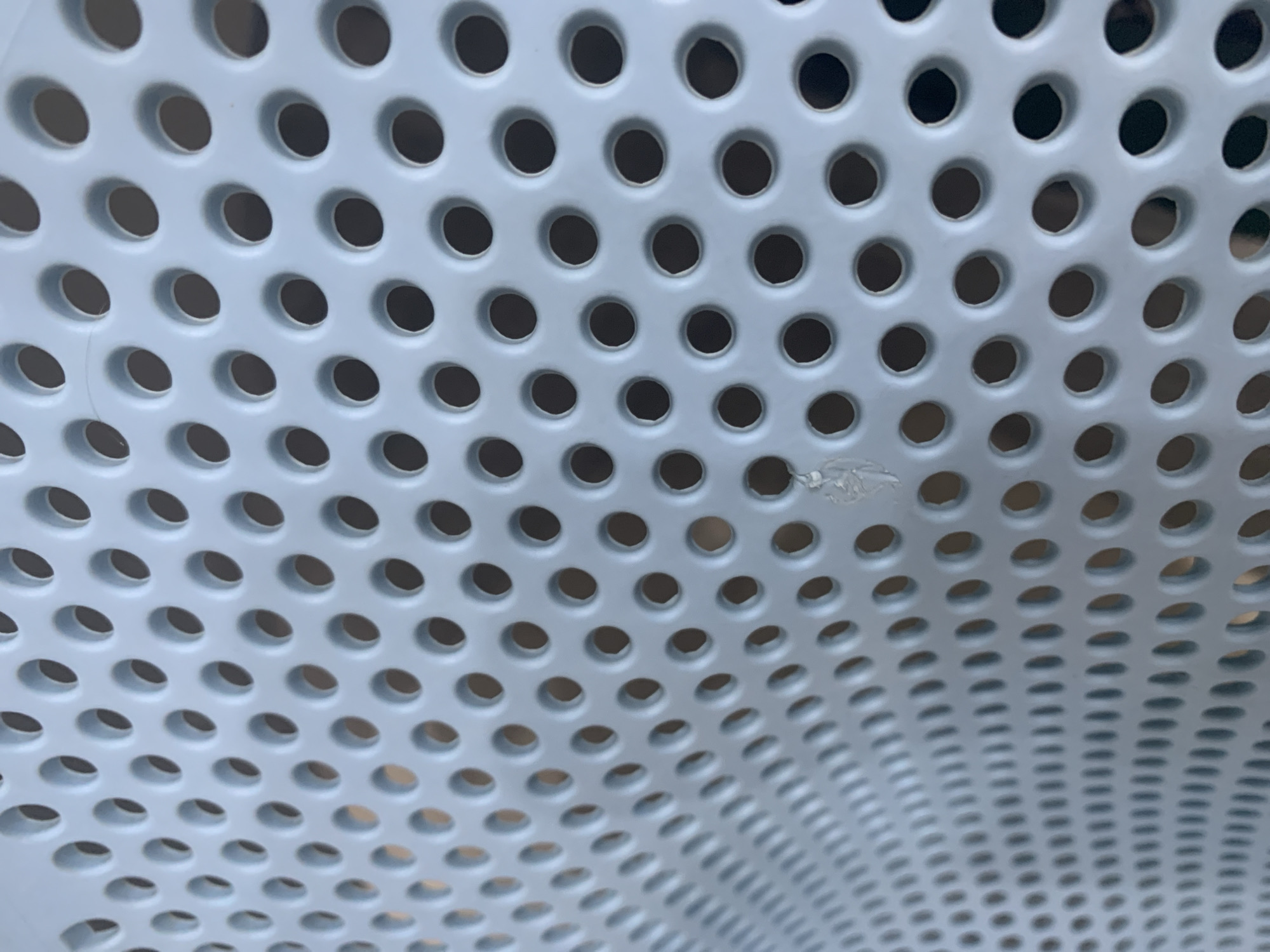 A close-up, high-angle shot shows a light blue, perforated surface, likely a metal chair or bench, with a repeating pattern of circular holes. The holes are of uniform size and are evenly spaced. The surface appears to be clean and slightly textured. In the center of the image, a small, light-colored speck of material is stuck to the surface, appearing as a slight discoloration and distortion of the holes in its immediate vicinity. The background is blurred and dark, suggesting the object is outdoors or in a dimly lit area. The composition and lighting emphasize the repetitive pattern of the holes. The image does not contain any elements that would indicate its location in Stavropol, Russia.