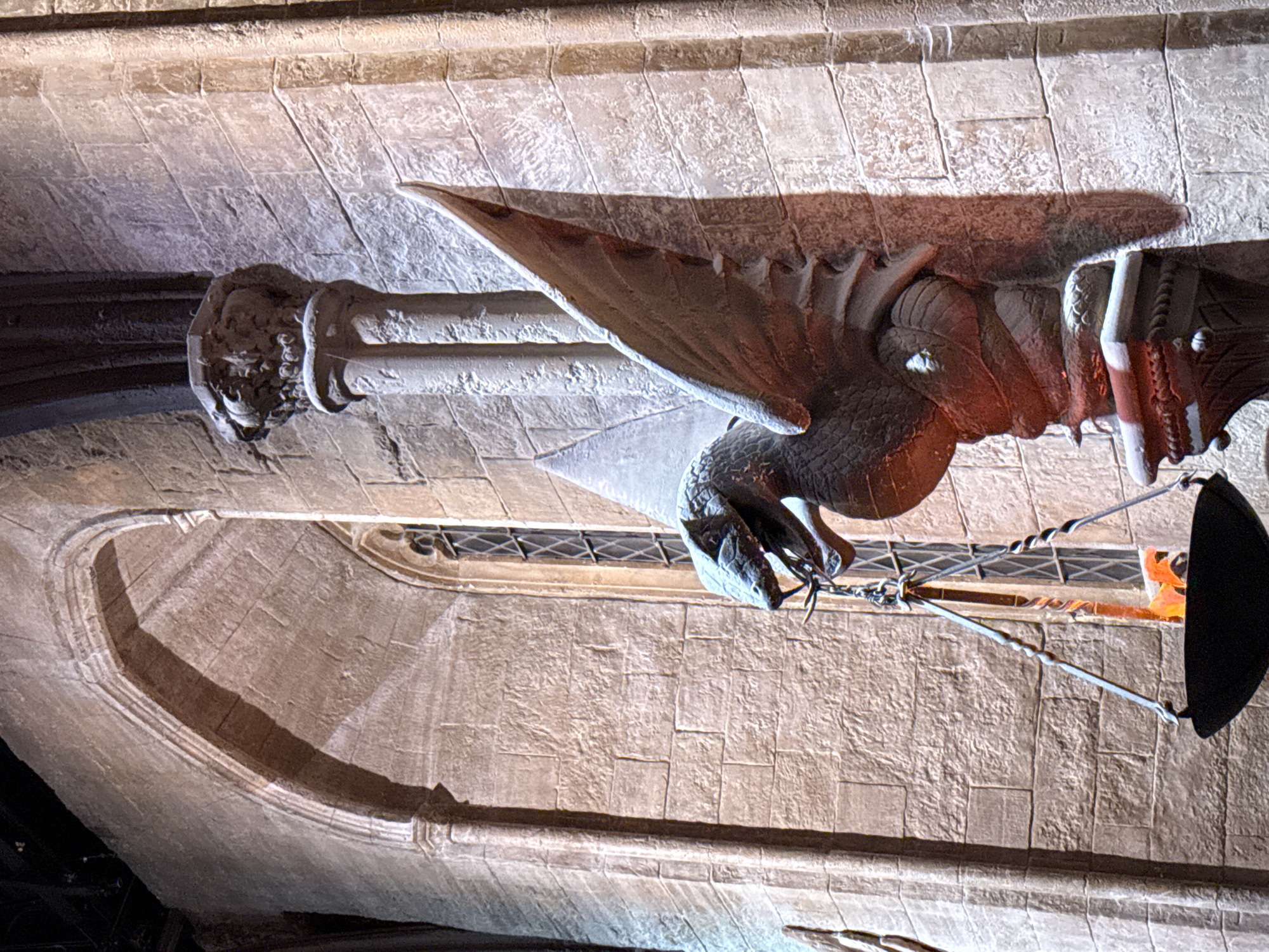 The image depicts a stone dragon sculpture with wings, coiled around a decorative pillar against a large stone wall. A hanging cauldron with flames is suspended from the dragon's mouth by chains. The architecture suggests a gothic or medieval style, with a pointed archway visible in the background and a tall, narrow window with a diamond-patterned grill. The lighting is dramatic, casting shadows and highlighting the textures of the stone. This scene is located within the Warner Bros. Studio Tour London - The Making of Harry Potter in Leavesden, Watford, United Kingdom, specifically within the Hogwarts Great Hall set. There are no people visible in the image.