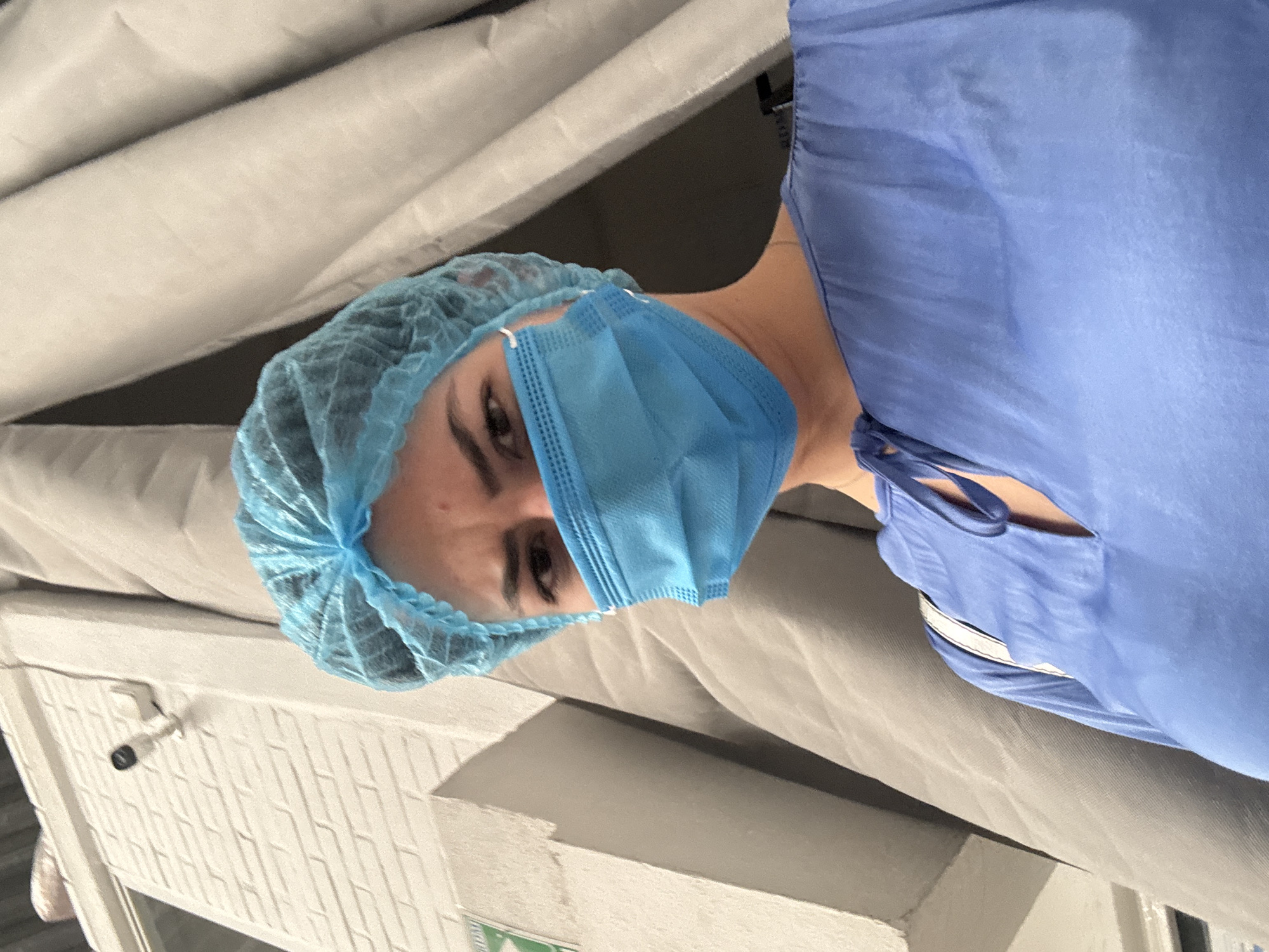 A person wearing a light blue surgical cap and a light blue surgical mask is depicted, facing slightly to the left of the frame. The person also has on a light blue surgical gown. The background consists of off-white folded material. In the bottom left corner, there is a white brick wall with a chrome lever and a green emergency exit sign. There is no information to suggest that the image was taken in the city of Puebla, Mexico.