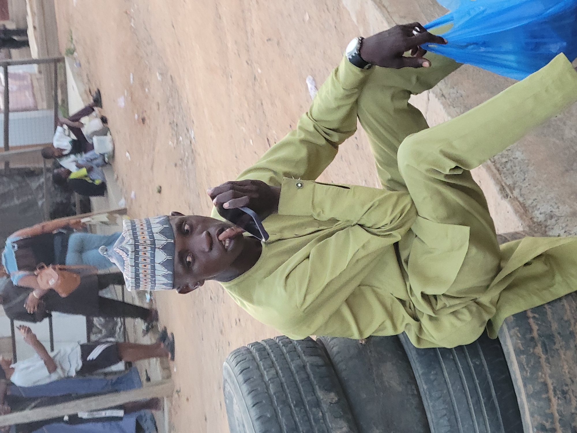 A man sits on a stack of tires in Suleja, Nigeria. He is wearing a light green traditional outfit and a patterned hat. He holds sunglasses in his right hand. He is also wearing a watch. To his right is a blue plastic bag. In the background, there are other people and buildings. The ground appears to be dirt or sand.