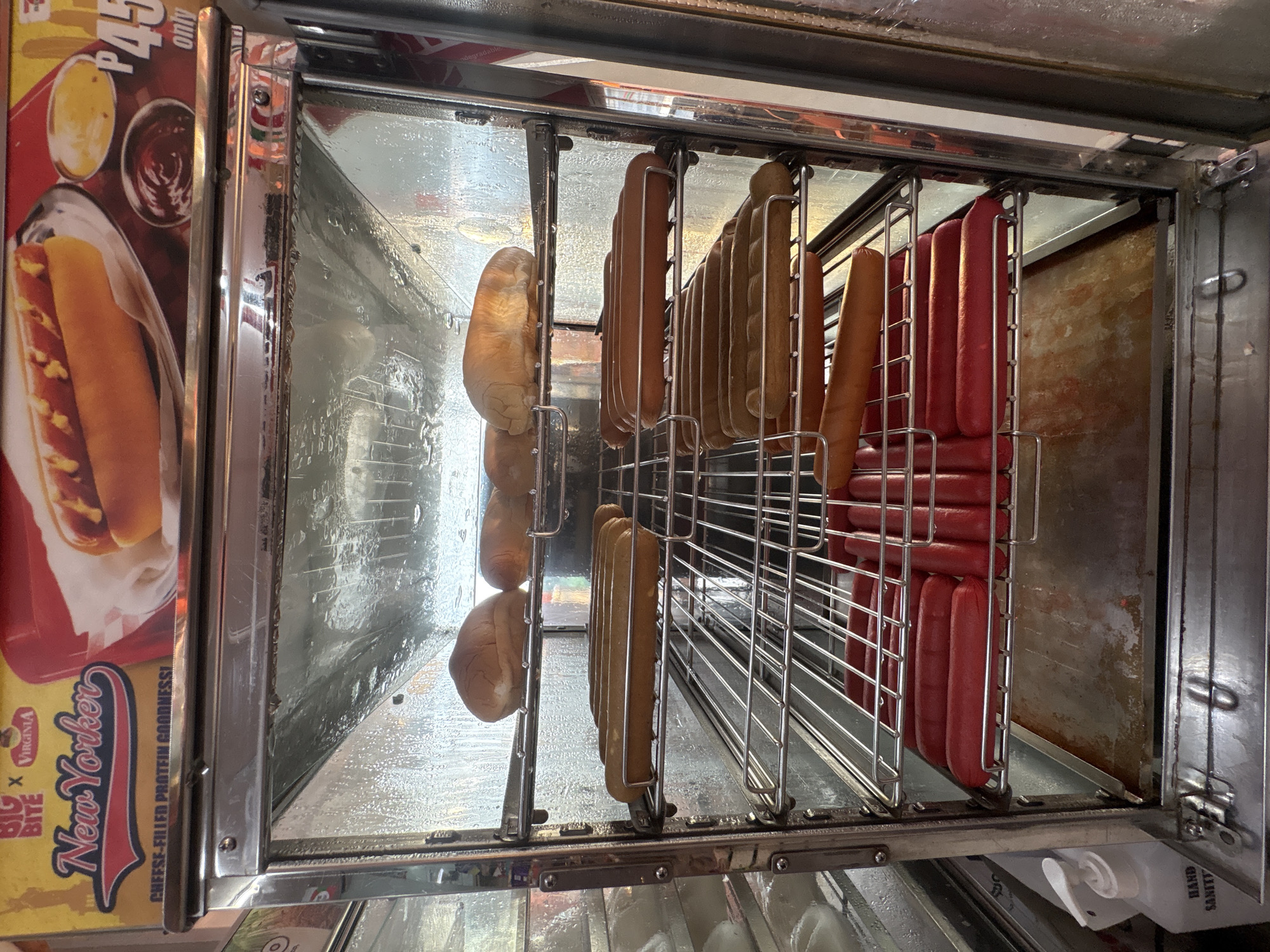 This is a medium shot, eye-level, indoor photograph of a hot dog warmer in Tagum City, Philippines. The warmer is made of stainless steel and glass and contains multiple shelves filled with hot dogs and buns. On the top shelf, there are three buns. The second shelf from the top has a rack with at least six hot dogs. The third shelf also has a rack with at least six hot dogs, which appear to be a different variety than the ones on the shelf above, as they are a lighter brown color. The fourth shelf from the top has a rack with several red hot dogs. There are also red hot dogs visible at the bottom of the warmer.
The top of the warmer displays a colorful sign advertising "BIG BITE New Yorker CHEESE-FILLED PROTEIN GOODNESS!" with an image of a hot dog on a bun with condiments. The sign also shows a price of "P45 only." The interior of the warmer appears to be steamy, suggesting the products inside are being heated.
To the left of the warmer, a white pump bottle of hand sanitizer is visible, with the label partially obscured but clearly showing the words "HAND SANITI." The lighting is bright, and the overall scene suggests a food service establishment.