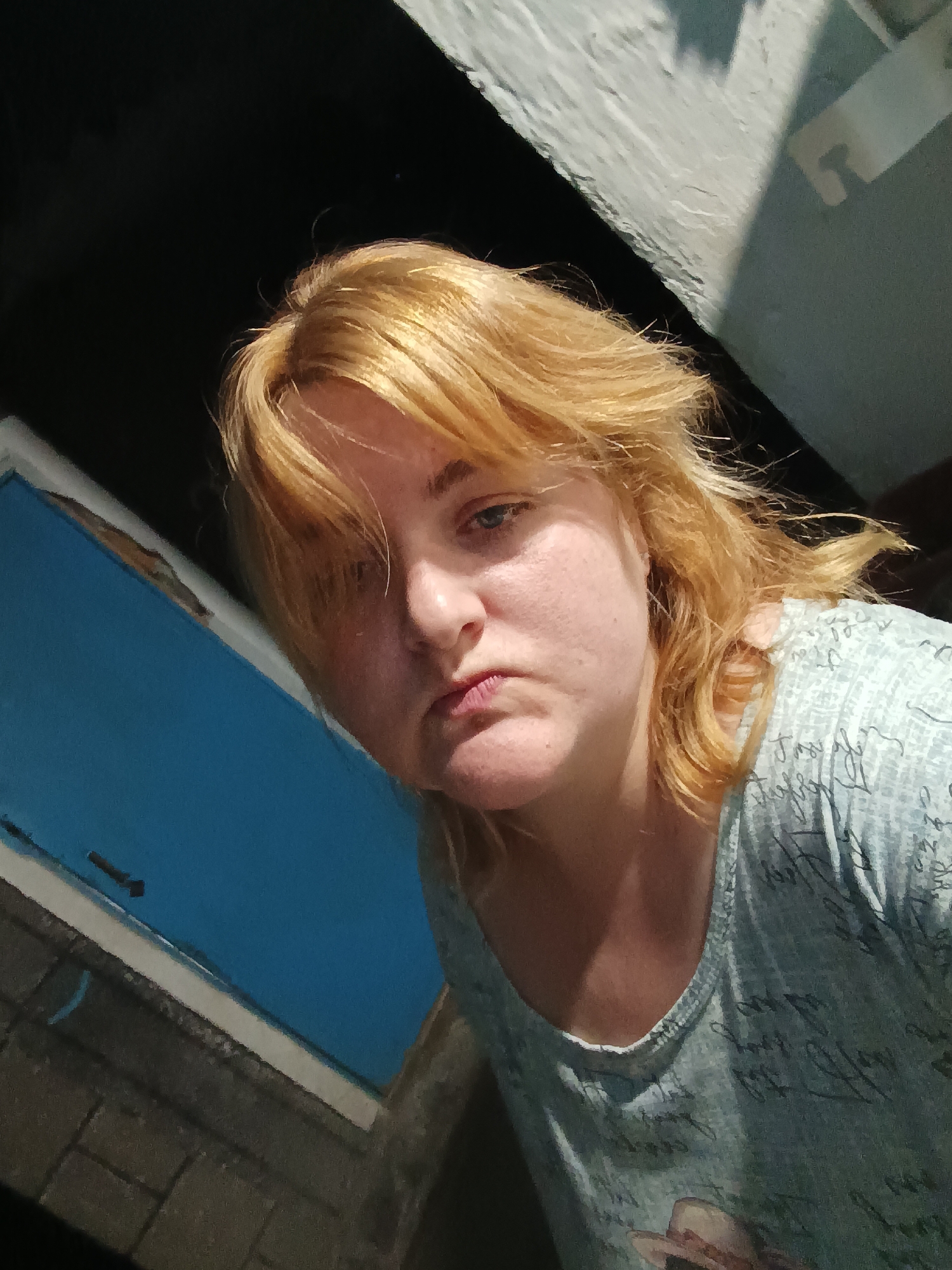A woman with blonde hair is shown in a close-up shot, looking down at the camera. She appears to be indoors, near a blue window frame. The background includes a dark ceiling and a textured wall. She is wearing a light blue patterned t-shirt. The location is Верхнесадовский округ, Ukraine.
