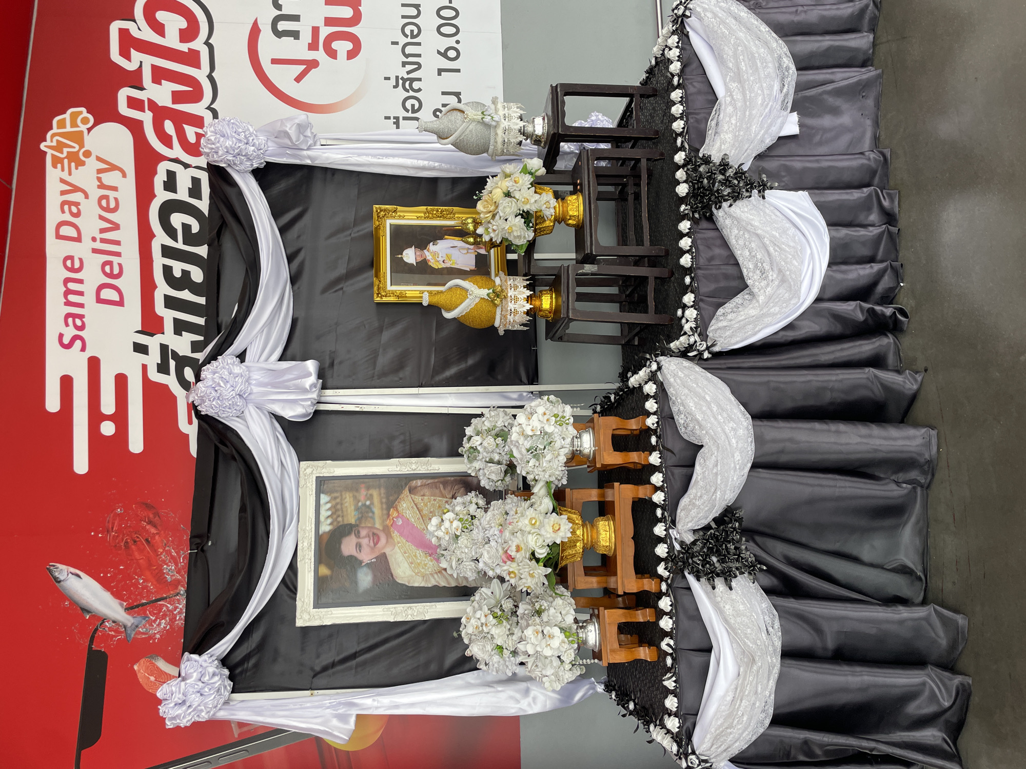 The image shows a memorial display set up outdoors, likely on a paved surface, in front of a red wall with commercial graphics. The display is draped in black and white fabric, with white floral arrangements and framed portraits. One portrait is of a woman in traditional Thai attire, and another is of a man in a military uniform. Two golden vases and decorative silver items are also part of the setup. The background graphics on the red wall include Thai text and images related to food delivery services. The overall presentation suggests a formal tribute.
