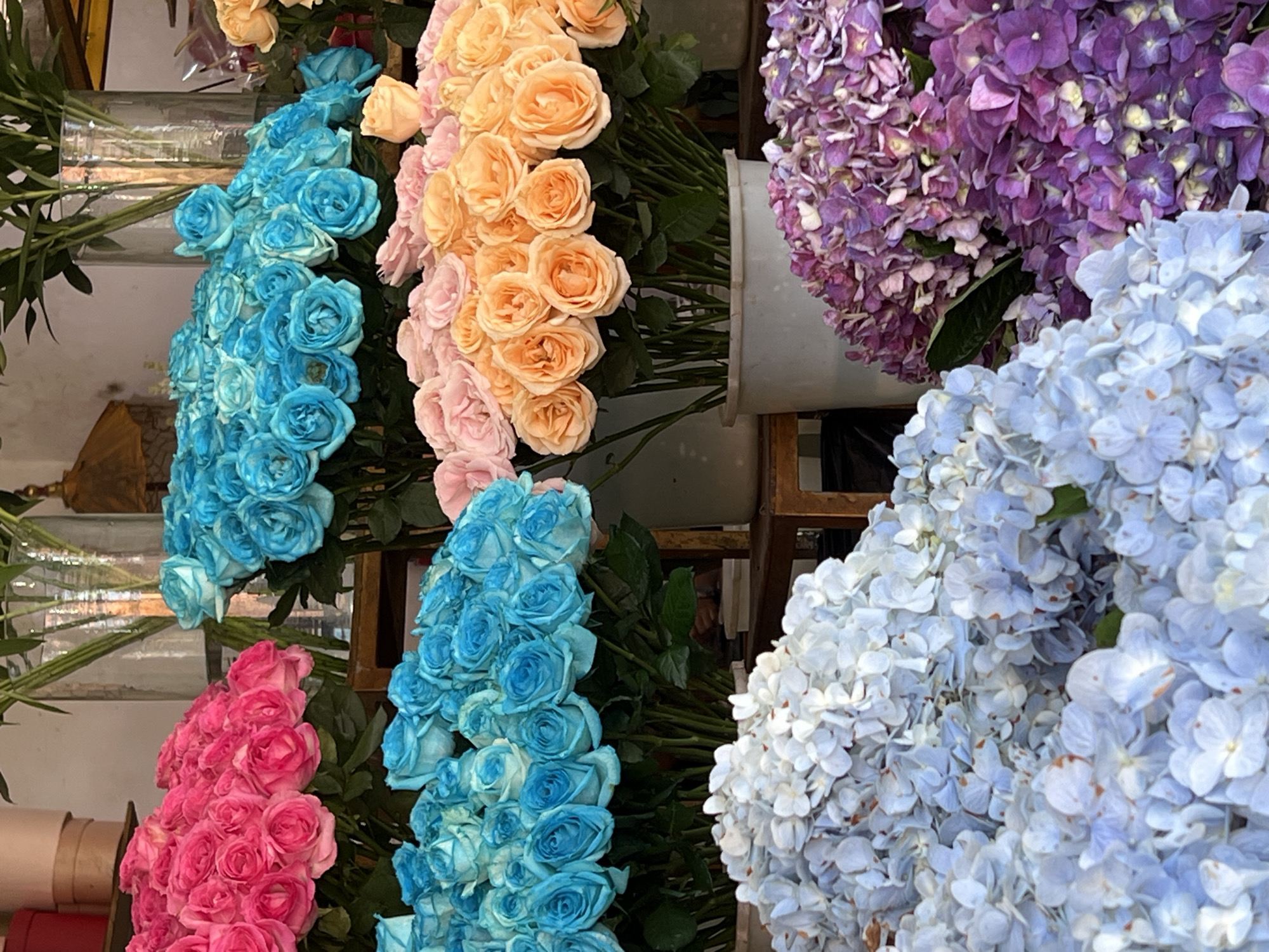 The image shows a display of flowers at a flower shop in Peliatan, Indonesia. Visible are several bunches of roses in various colors including pink, blue, and peach. There are also bunches of purple and light blue hydrangeas. The flowers are arranged in containers and likely on shelves or a stand within the shop. The lighting is bright, suggesting it's either an indoor space or an outdoor setting with ample natural light.