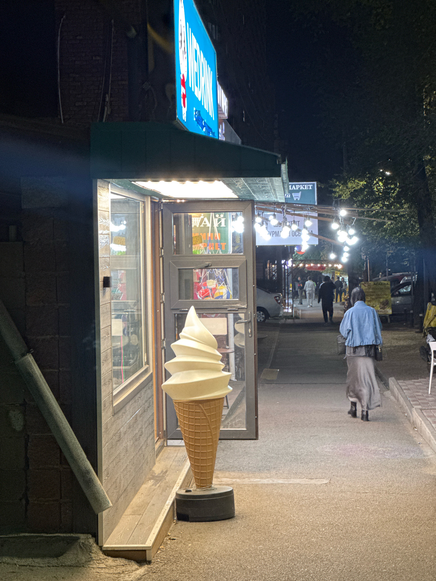 This is a nighttime, eye-level, medium shot of a street scene in Almaty, Kazakhstan, featuring an ice cream shop entrance. The shop, to the left, has a large, illuminated blue sign above it with white lettering that appears to say "WEBRINK". A prominent, oversized sculpture of a vanilla soft-serve ice cream cone stands outside the entrance. The shop's interior is visible through its glass doors and windows, showing various items for sale.
The street is paved and lined with trees on the right. Several people are walking along the street in the background, some illuminated by string lights that hang overhead. To the right of the ice cream shop, a sign reads "LAPKET" in white lettering on a dark background, with a shopping cart icon. Further down the street, there are cars parked and a few more people are visible. The overall atmosphere is that of a lively commercial street at night.