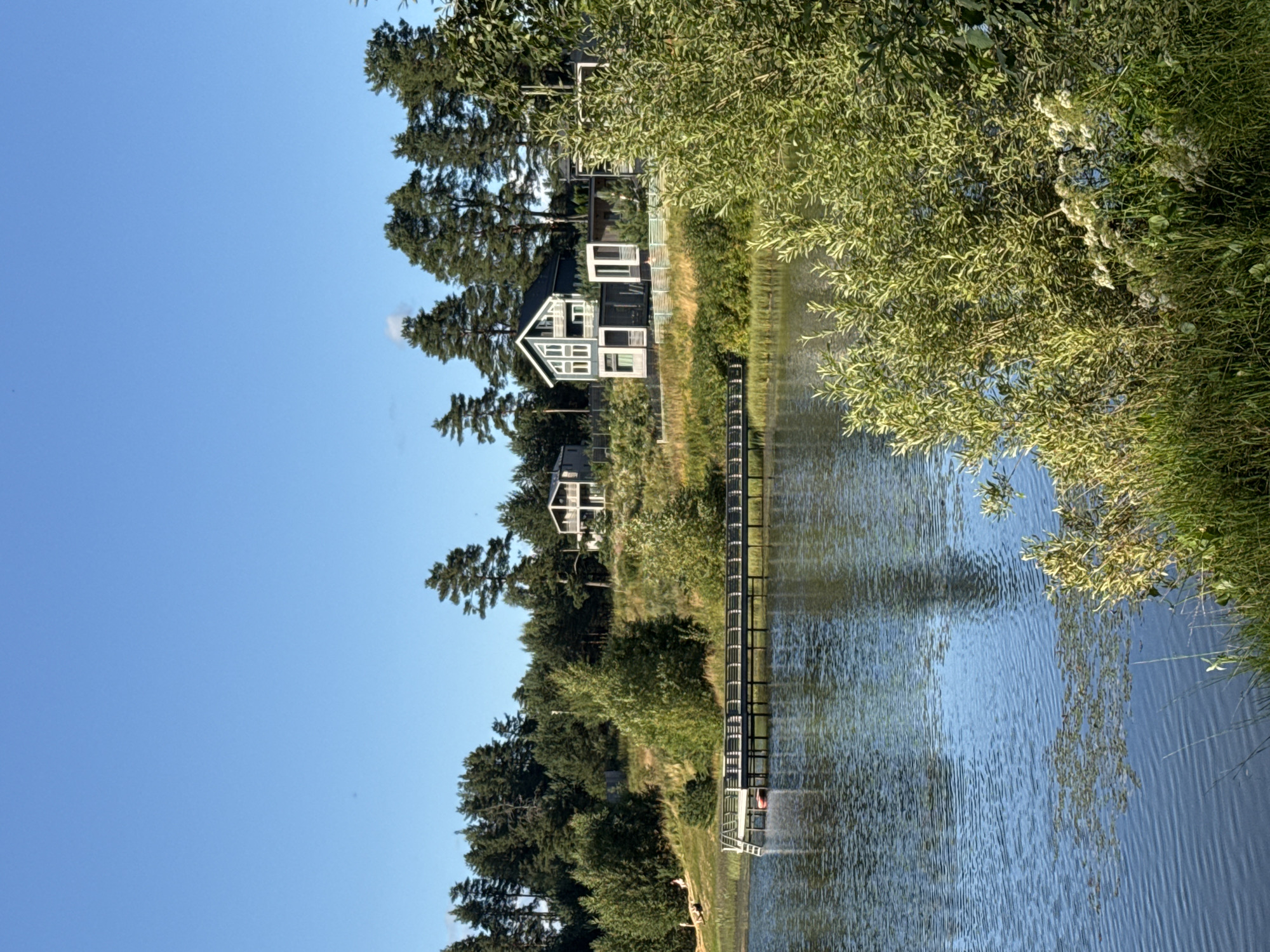 The image shows a serene lakeside landscape. In the foreground, the tranquil, blue water of a lake reflects the clear sky and surrounding vegetation. Some aquatic plants are visible on the surface of the water. The middle ground features a wooden pier extending into the lake, with a small red object, possibly a boat or buoy, attached to it. The background is dominated by a lush, green hillside covered in trees, with a few modern houses nestled amongst them. The houses have contemporary architecture with large windows and balconies. The sky above is a vast expanse of bright, cloudless blue. A few people are casually seated on the grassy slope to the left of the pier. The overall impression is one of a peaceful, semi-rural setting. The location is not specified as being in the city of Mars, Russia.