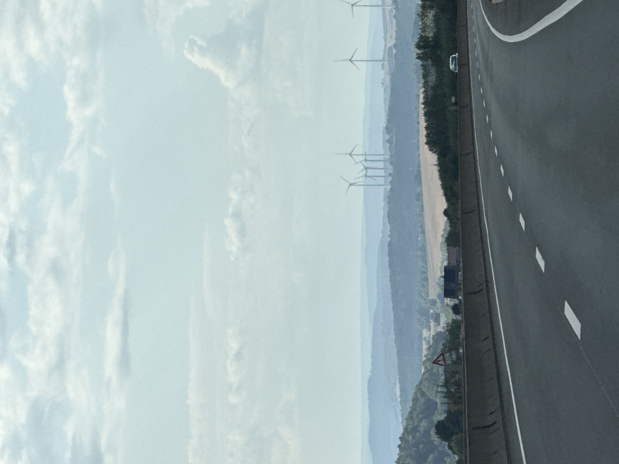 The image captures a view from a highway, looking towards rolling hills in the distance under a cloudy sky. Several wind turbines are visible on the horizon, silhouetted against the sky. In the foreground, the asphalt road is marked with white lane lines. A concrete barrier separates the road from the roadside. On the side of the road, there's a triangular warning sign. Further in the distance, beyond the trees and fields, a white car is visible on the highway. The overall scene suggests a rural or semi-rural landscape, likely a highway passing through a hilly region with wind farms.