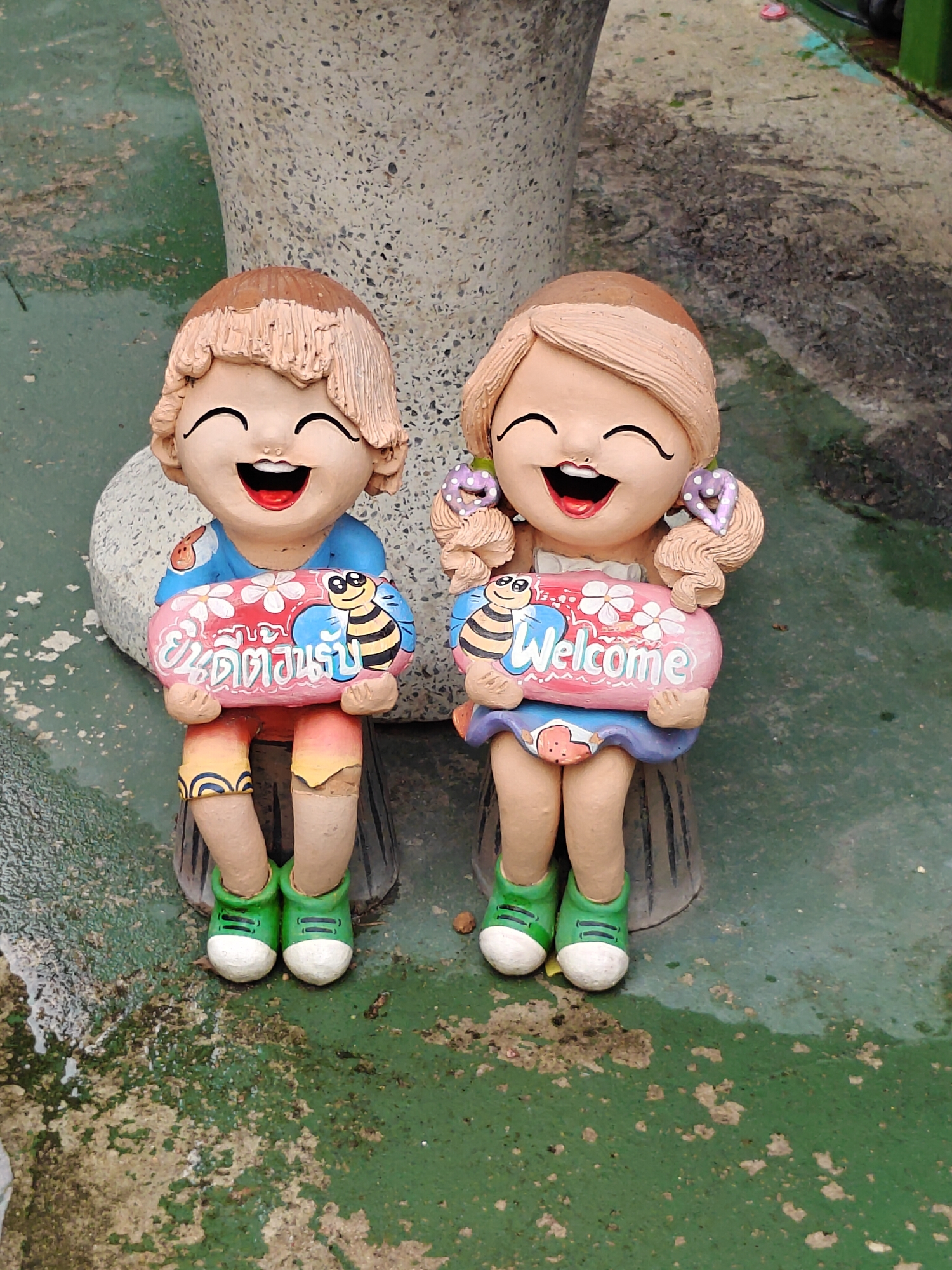 Two ceramic garden gnomes, a boy and a girl, are seated on a weathered green surface that appears to be a paved area with some patches of discoloration and moisture. The gnome on the left is wearing a blue shirt and orange shorts, and holds a pink sign with Thai writing and a bee. The gnome on the right wears a purple skirt and holds a pink sign with the word "Welcome" and a bee. Both gnomes have wide smiles and are dressed in green sneakers. Behind the gnomes is a large, rough-textured, grey concrete object, possibly part of a planter or a statue. The overall scene suggests an outdoor decorative element in a garden or public space.