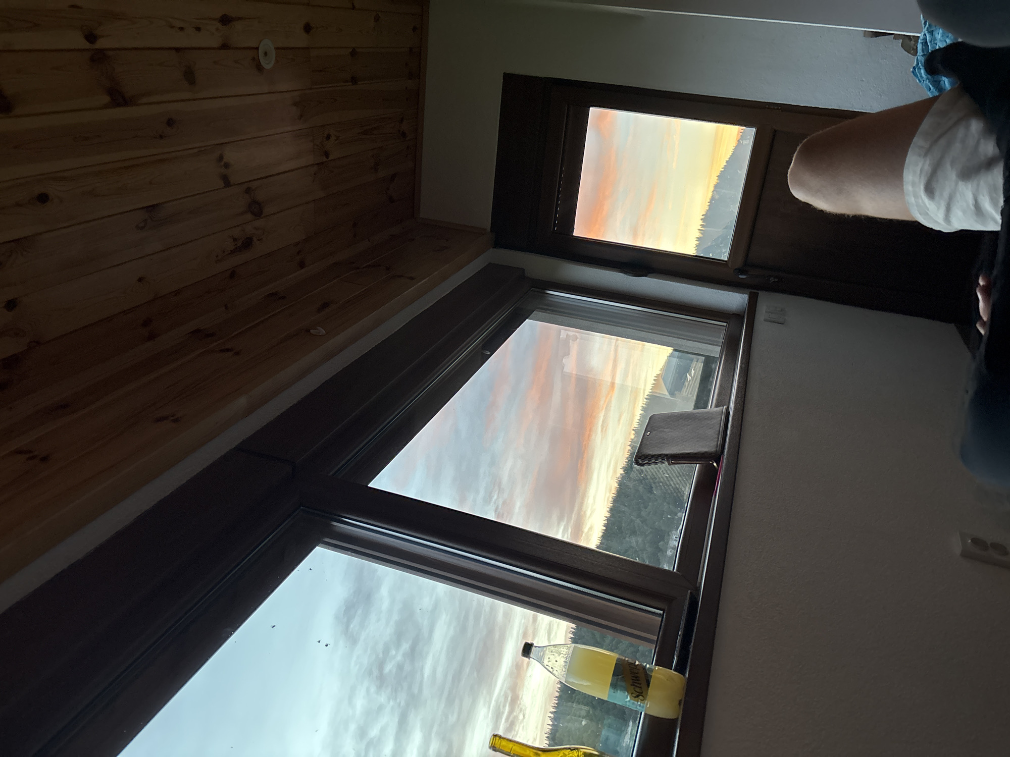The image captures a serene view of a sunset over a forested landscape, observed from inside a room with a wooden paneled ceiling. Large windows offer a panoramic vista, with the sky painted in hues of orange, pink, and blue. On the windowsill of the larger window, a bottle of "Schweppes" sparkling lemonade is placed next to a yellow bottle. A dark, rectangular object, possibly a book or a wallet, rests on the sill as well. To the right, a smaller window or doorframe reveals a similar sunset view. The lower portion of the image shows a person's leg, clad in white shorts, resting on what appears to be a bed. The overall ambiance suggests a moment of relaxation and appreciation of nature's beauty. This scene is not located in Virak, Montenegro.