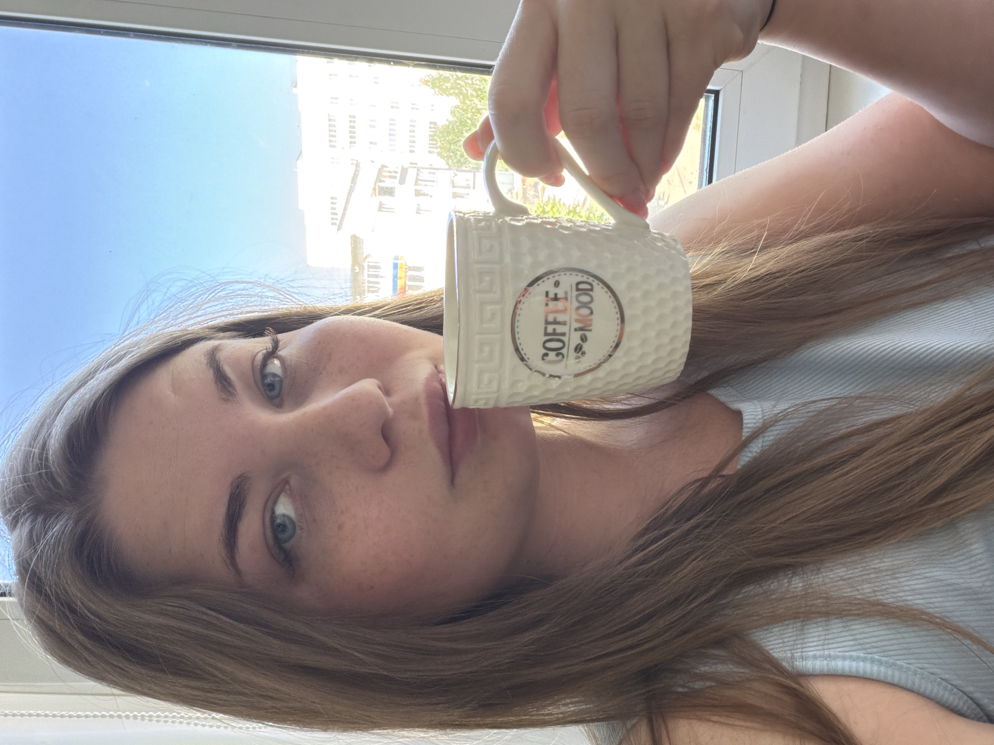 A woman is holding a white coffee mug with the words "Coffee mood" on it up to her lips. She has light brown hair, blue eyes, and freckles. She is wearing a light blue shirt. The background shows a building and a blue sky visible through a window. The location is Chaikovsky, Russia.