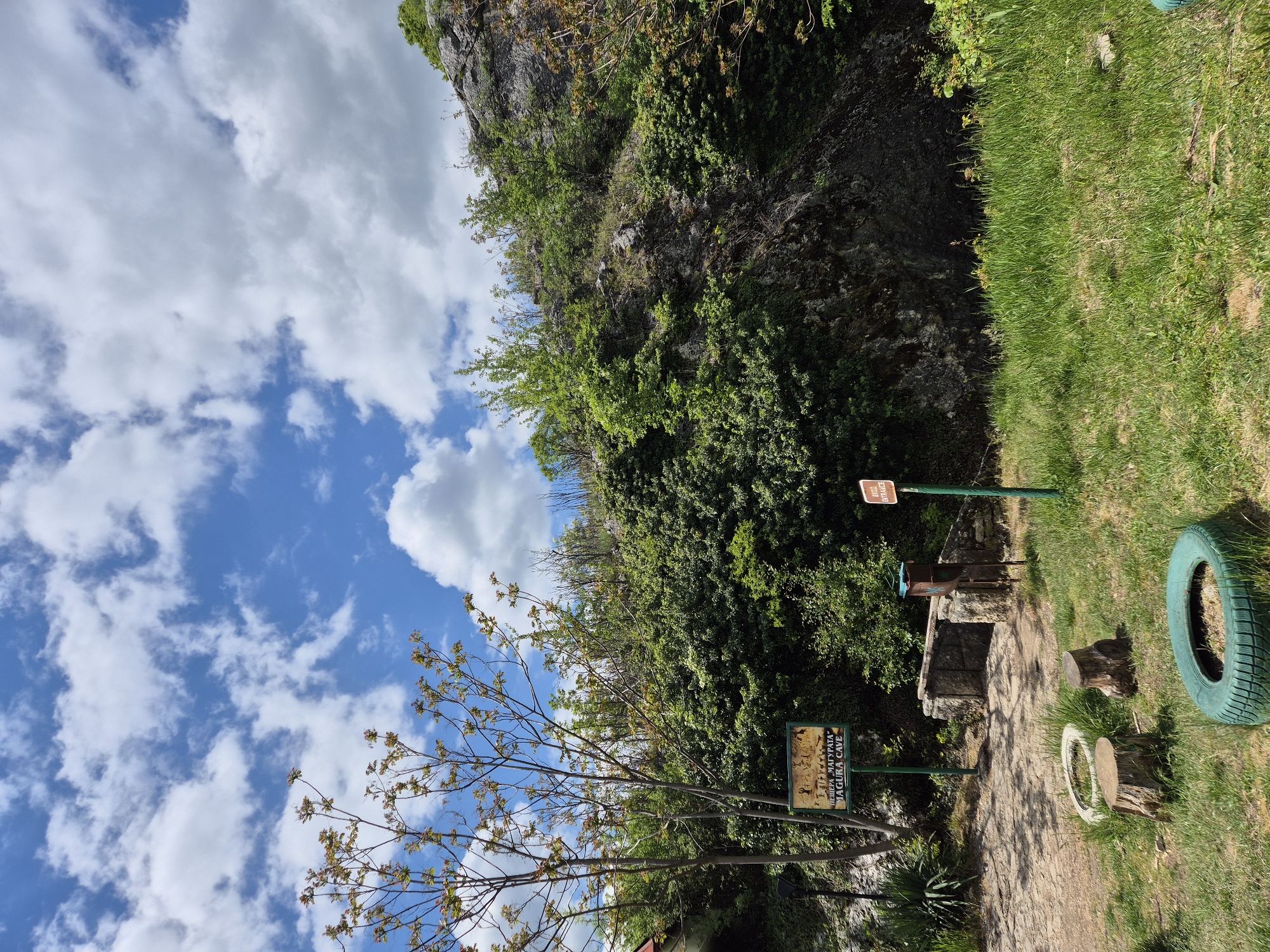 The image depicts a pathway in Raianovtsi, Bulgaria, leading toward the entrance to the Magura Cave. A rocky cliff face covered in vegetation is visible to the right of the path. A sign with text in Cyrillic and English ("Magura Cave") is present alongside the path. The sky is blue with scattered clouds, visible above the trees. A green-painted tire sits on the grass beside the path. A sign that says "Cave Entrance" sits above the entrance to the cave.