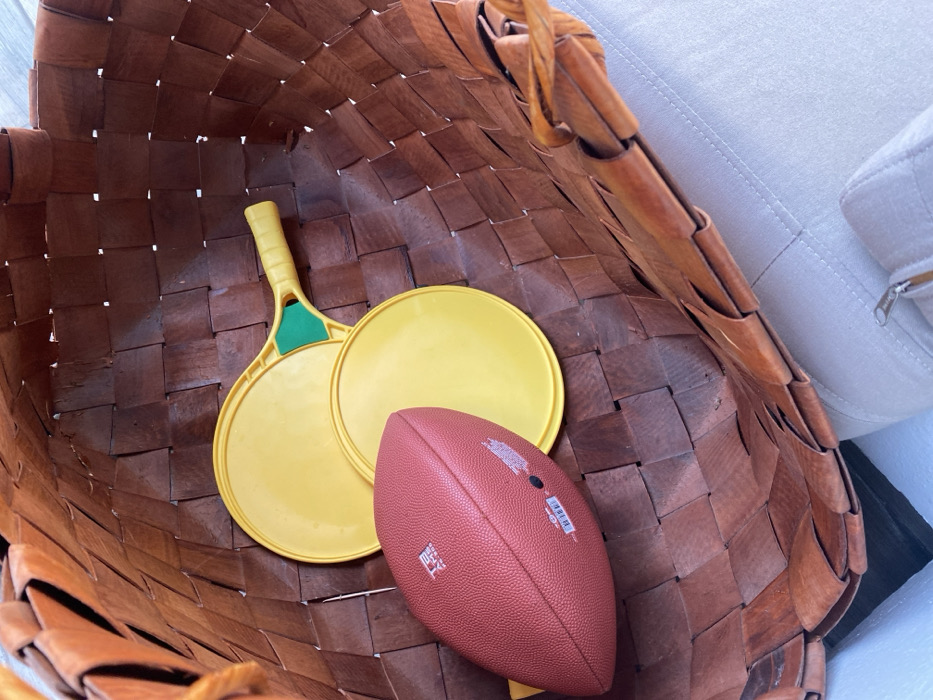 Inside a woven, brown basket are two yellow beach paddle ball rackets and a red-brown American football. The rackets are overlapping each other. The football lies near the paddles, slightly covering one. The basket's weave pattern is visible, adding texture. A portion of a light-colored, upholstered couch is visible in the upper right corner.