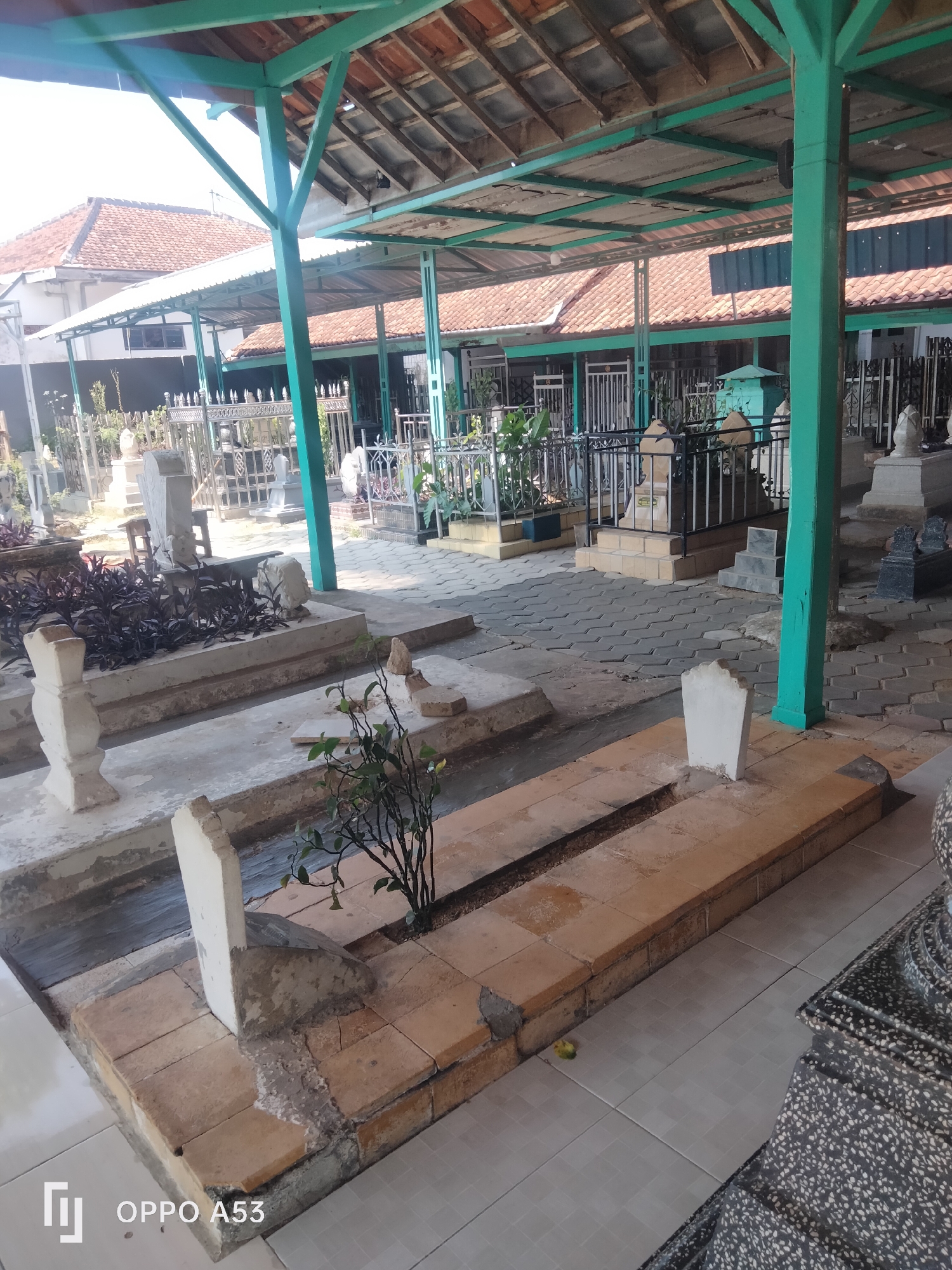 The image depicts a cemetery with multiple graves, some covered by a green-painted metal awning with a corrugated roof. The graves are of various designs, with some having white stone markers and others being simple elongated mounds. The ground is paved with hexagonal tiles in some areas and with rectangular tiles in others. There are also some plants and bushes present within the cemetery. In the background, there are buildings with red tile roofs. The overall scene suggests a burial ground in a tropical climate, possibly in a residential area given the proximity of the buildings.