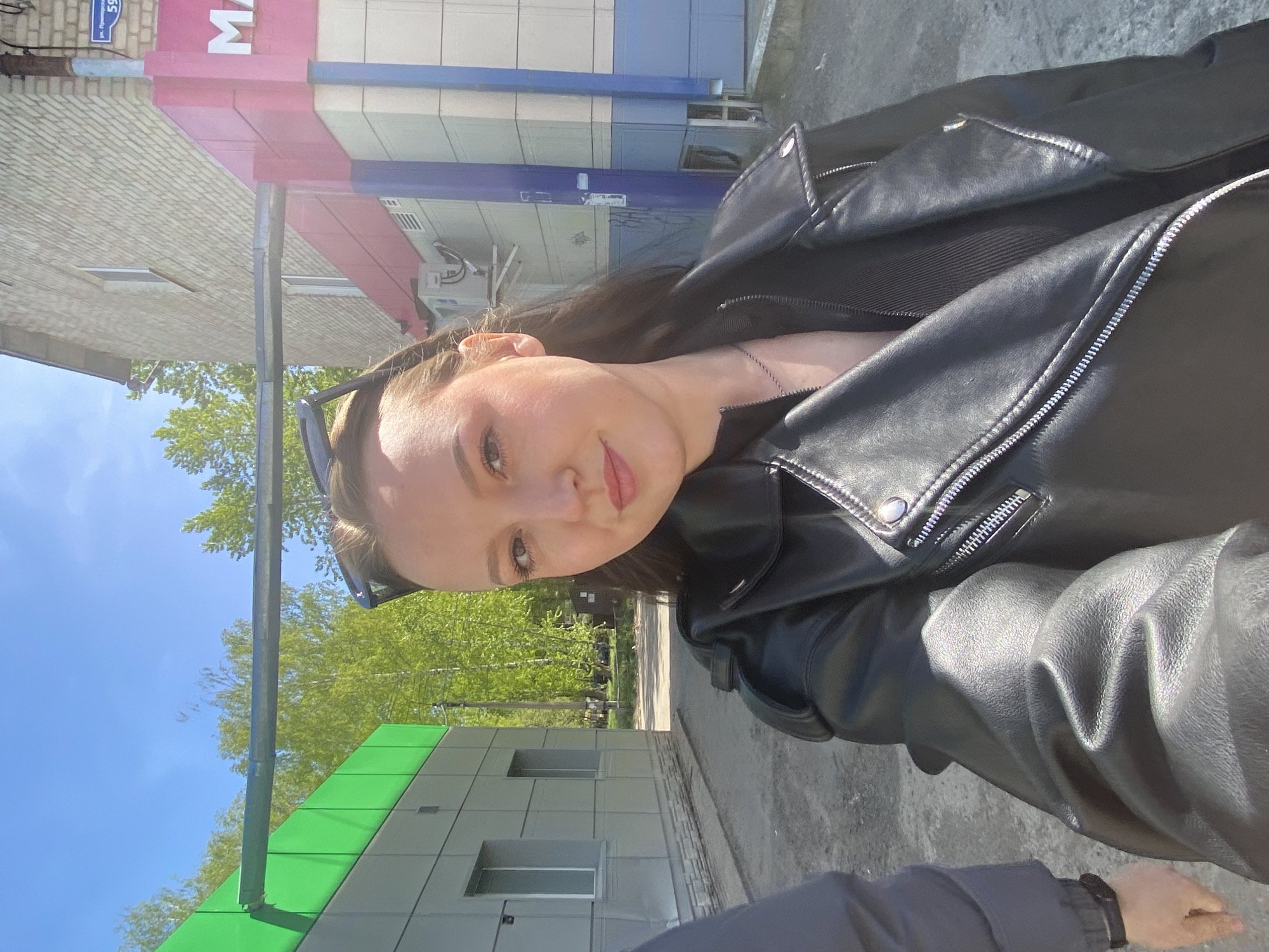 A young woman, seen from the chest up, is outdoors in what appears to be a city environment, potentially in Chaikovsky, Russia. She is wearing a black leather jacket over a black top and a silver chain necklace. Sunglasses are perched on her head.
The background shows parts of buildings. One building features a pink and white facade with the partially visible letter "M". Another building has a green and gray exterior with visible windows. Trees are visible in the distance against a blue sky. The ground appears to be paved.
