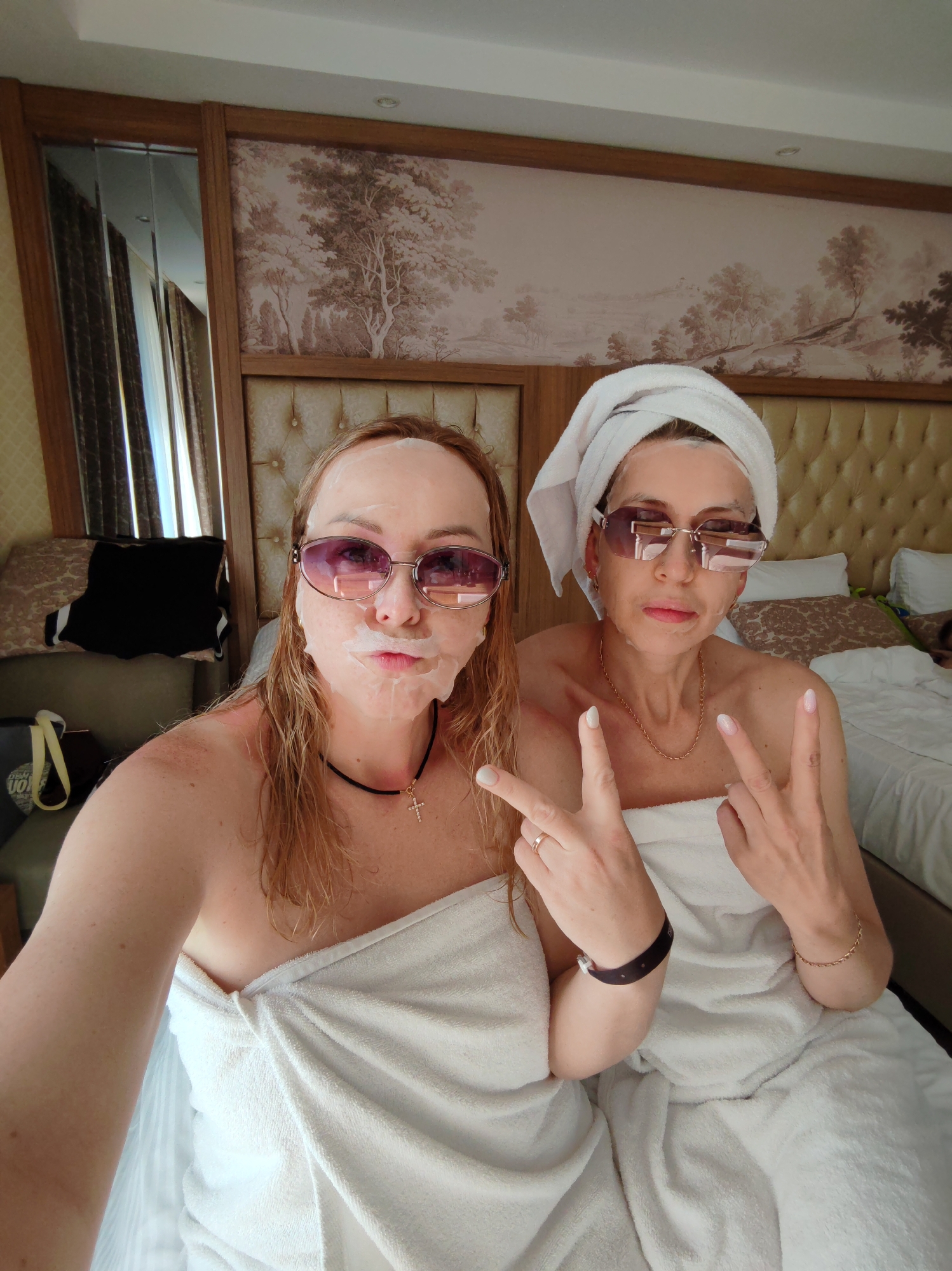 Two women, wearing white towels and sunglasses, are pictured indoors. The woman on the left has wet hair and a white facial mask, adorned with a black choker necklace. The woman on the right has her hair wrapped in a white towel and wears a gold chain necklace. Both are making a peace sign with their hands. The room features a padded headboard, decorative wallpaper, a mirror, and a bed with white bedding. The location is likely a hotel room in Alanya, Turkey.