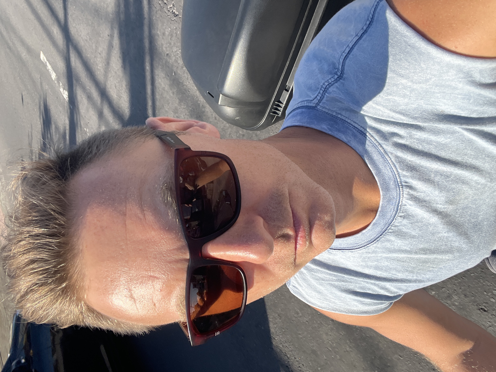 A man wearing sunglasses and a light blue sleeveless t-shirt is captured in a selfie-style shot outdoors. The man has short, blondish-brown hair and his facial features, including his nose and lips, are visible. His sunglasses have dark, reflective lenses. In the background, a dark object, possibly a motorcycle saddlebag, is present. The ground appears to be asphalt, with some shadows and a white line visible, suggesting a road surface. The lighting indicates it is daytime with bright sunlight. The scene does not provide any specific visual cues to determine the location within Sukawati, Indonesia.