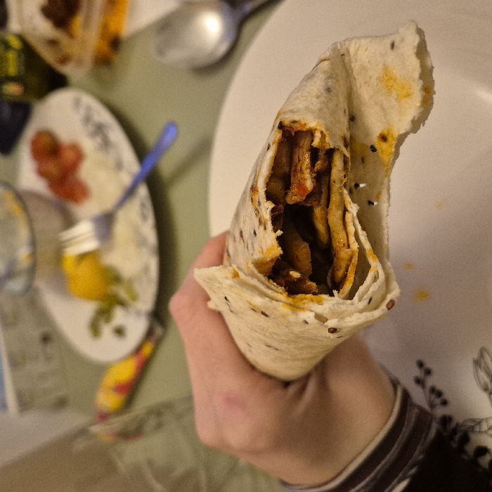 A hand holds an open tortilla wrap, revealing its filling of meat strips and sauce. The wrap appears to be a flatbread with speckles throughout. A plate with tomatoes, white dressing, and yellow dressing is visible in the background, along with a spoon and another plate with a partial wrap. The setting appears to be a casual dining environment. The person's wrist shows a striped sleeve.