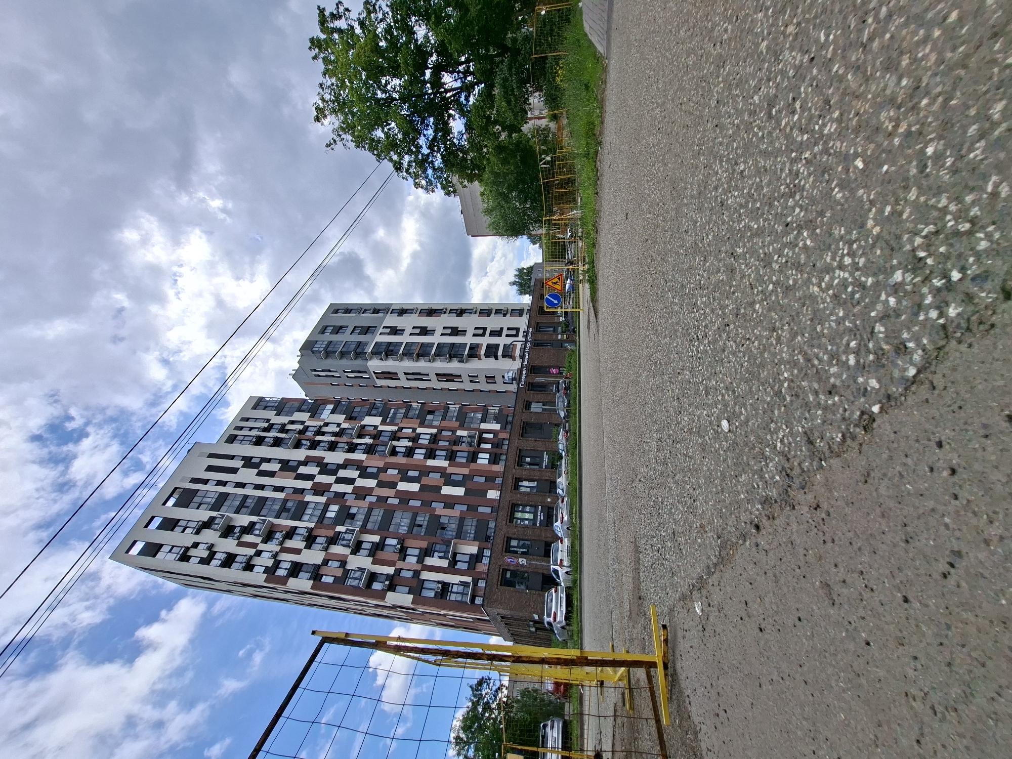 This is a low-angle, wide shot of a street scene in Izhevsk, Russia. The foreground is dominated by a textured asphalt road. To the left, a modern apartment building with a distinctive brown and white geometric facade rises high into the sky. Behind it, another building with a lighter facade is visible. A yellow temporary fence with a net is in the lower left corner. Overhead, power lines stretch across a cloudy sky with patches of blue. Green trees are visible behind the fence and to the right of the buildings. A few cars are parked along the street in front of the buildings, and some traffic signs, including a yellow triangular sign with a black symbol and a blue circular sign with a white arrow, are visible near the road. The overall impression is an urban environment on a day with mixed weather. The time of day appears to be daytime, given the natural light. No people are visible in the image. A partially visible sign on one of the buildings has Cyrillic text, possibly indicating a business.