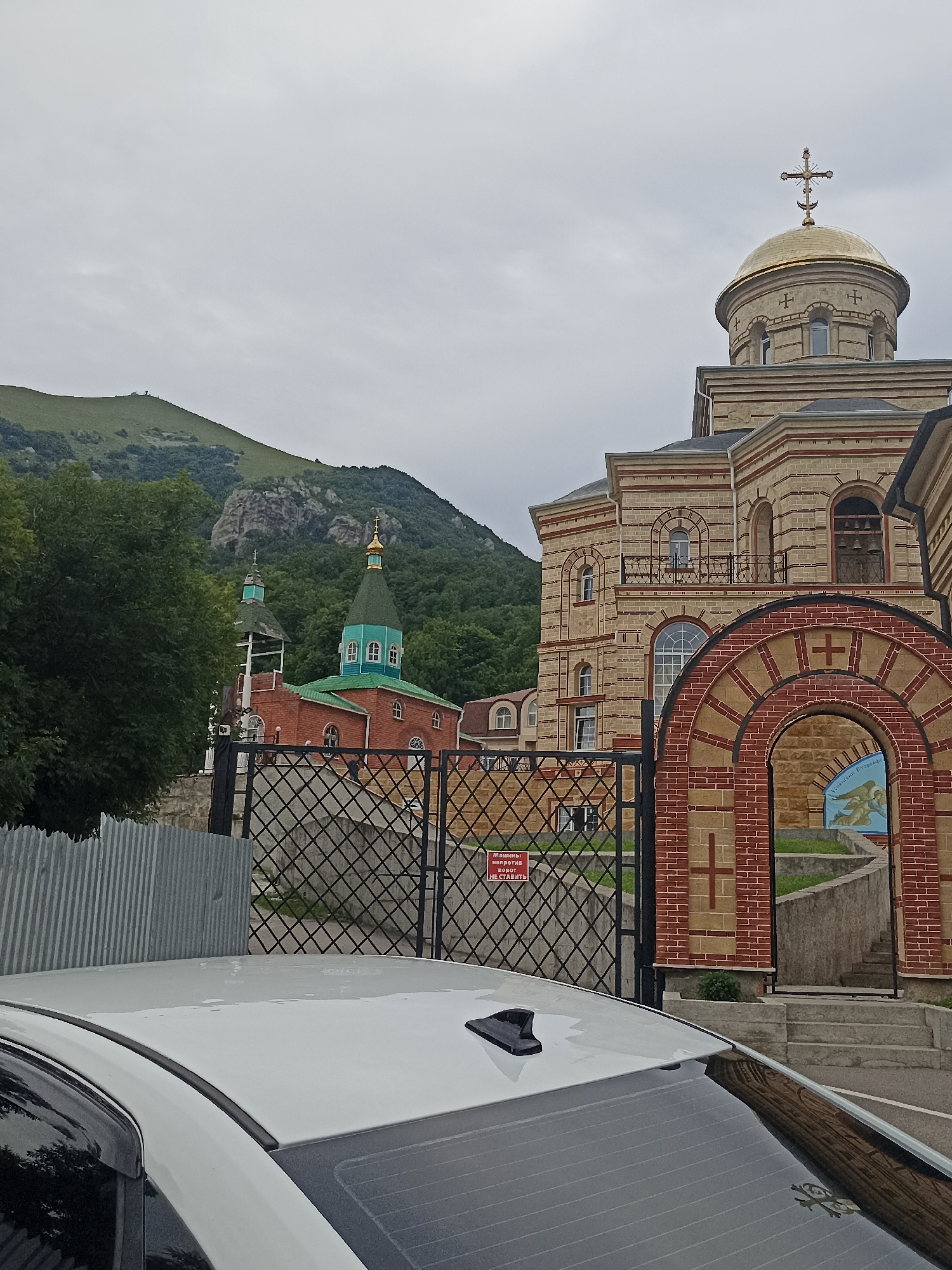 The image captures a view of the Church of the Intercession of the Most Holy Theotokos in the city of Lermontov, Russia. The church features a prominent golden dome topped with a cross, constructed from light-colored stone with red brick accents. To the left of this main structure, a smaller red brick church with a green dome and spire is visible, nestled amidst lush green trees. In the background, a sloping, green mountain range under a cloudy sky dominates the landscape. In the foreground, the white roof of a car is visible, along with a metal fence and a gate. A red sign with white lettering is affixed to the gate.