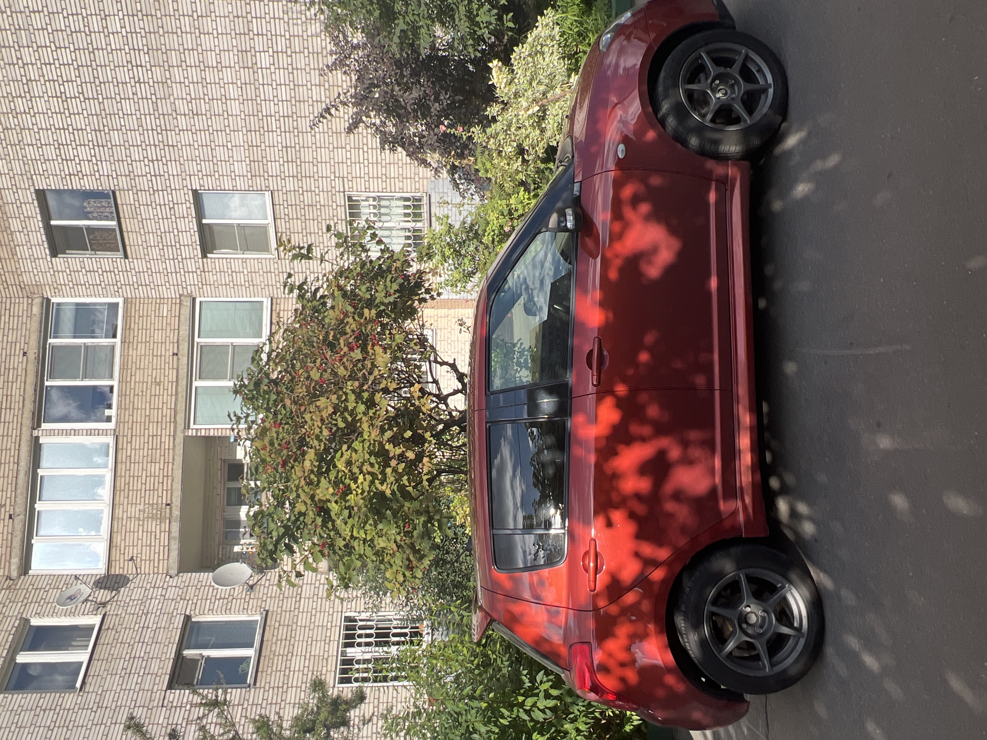 A red Suzuki Swift is parked on asphalt in front of a pale brick apartment building. The car is viewed from the side, with its rear on the left and its front on the right. The building has multiple floors and windows, some of which are partially covered by white curtains. Several satellite dishes are attached to the exterior of the building. Green trees and bushes are growing around the building and partially obscure the lower floors. Sunlight casts dappled shadows of leaves onto the car and the ground. The image was taken on Naberezhnaya Ulitsa in Khimki, Russia.