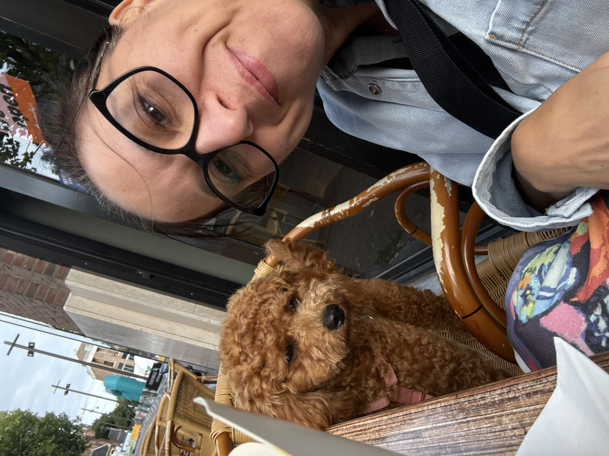 A woman with dark hair and glasses is taking a selfie with her brown poodle. They are sitting at a table outside a cafe, possibly in Groningen, Netherlands, as indicated by the street scene in the background. The poodle is sitting in a wicker chair next to the woman. The woman is wearing a light blue denim jacket. The cafe appears to be on a street with buildings and trees.