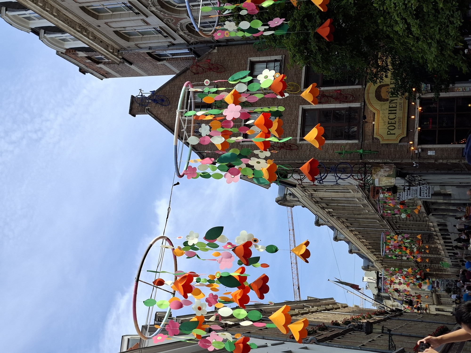 This image captures a street scene in Brussels, Belgium, specifically in the Rue de Remparts, a charming street lined with historic buildings. The street is adorned with festive decorations, with colorful paper flowers and leaves suspended from large circular frames, creating a vibrant, whimsical atmosphere. These decorations are hung above the street, casting a cheerful ambiance.

In the background, traditional Brussels architecture dominates, with multi-story brick buildings featuring arched windows and ornate facades. Some buildings display signage, including one for "POCHENELLE ESTAMINET - LEGEND OF BEER", hinting at the culinary and cultural offerings of the area. Decorative bicycles are also visible on the sides of some buildings, adding a unique touch to the urban landscape.

The street is bustling with people, who appear to be enjoying a leisurely stroll. Some individuals are seen taking photographs, capturing the lively scene. The sky above is a clear blue with scattered clouds, suggesting a pleasant day. A construction crane is faintly visible in the distance, a subtle reminder of ongoing urban development amidst the historic charm. The overall impression is one of a vibrant, culturally rich European city street, decorated for a celebration or festival.