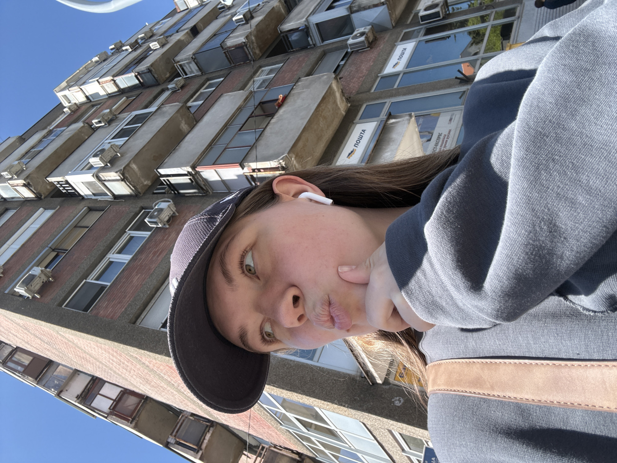 A woman in a gray sweatshirt and a black baseball cap is posing for a selfie. She is wearing an earbud in her left ear and has a hand under her chin. Behind her is a tall building with multiple windows and air conditioning units. The windows have signage on them written in Cyrillic. The building is in the Vozdovac Urban Municipality, Serbia. The sky is clear and blue.