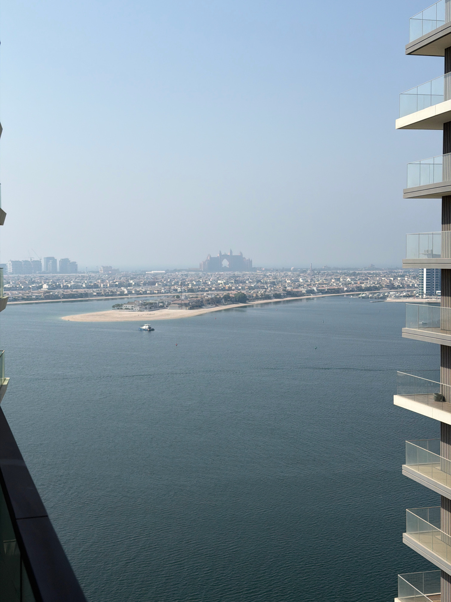 This is an aerial view looking towards the Palm Jumeirah in Dubai, United Arab Emirates. In the foreground, the edge of a modern residential building with glass balconies is visible on the right side of the frame. The middle ground features the distinctive fronds of the Palm Jumeirah, a man-made archipelago, with a calm, dark blue body of water in front of it. A small white boat is visible on the water near the sandy shore of one of the fronds. Beyond the Palm, a sprawling cityscape stretches across the horizon, characterized by numerous buildings, including some taller structures and cranes, indicating ongoing development. In the far distance, a hazy outline of a larger, prominent building with arched structures is visible, likely the Atlantis, The Palm hotel. The sky is a clear, pale blue with a slight haze, typical of the Dubai climate. There are no people visible in the frame.