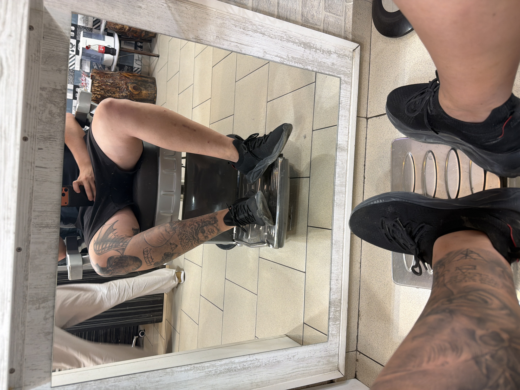 The image shows a person's legs and feet, reflected in a mirror. The person is wearing black sneakers and black shorts. Their legs are covered in tattoos. They are sitting in a barber's chair, which is visible in the reflection. The floor is tiled with light-colored tiles. In the reflection, another person is visible wearing white pants. The background of the reflection shows a barbershop interior with wooden decorative elements, a cooler, and a Coca-Cola bottle. The mirror frame is white with a distressed wood-like texture. The setting appears to be a barbershop in Ruse, Bulgaria, given the provided context.